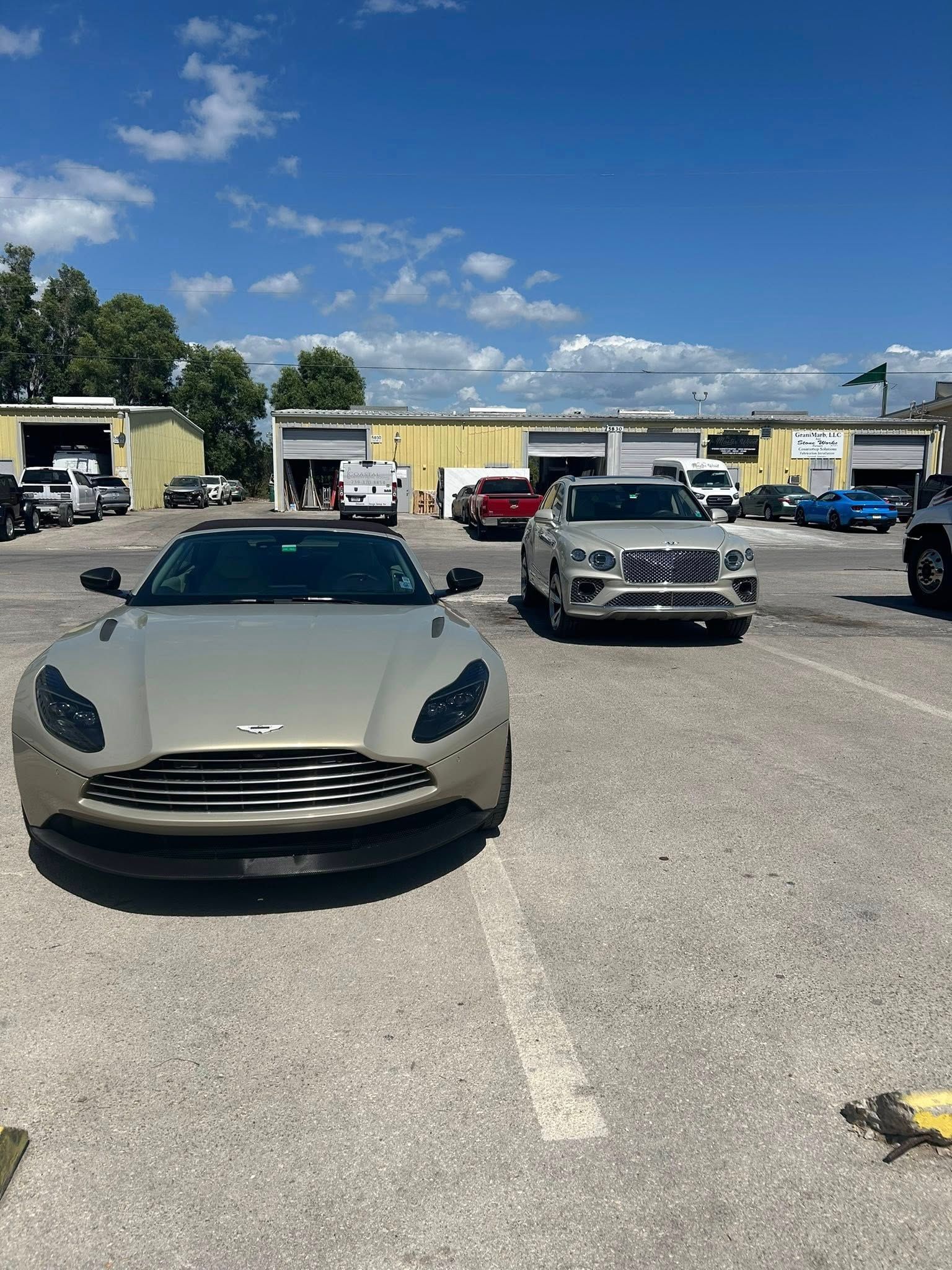 A taupe Aston Martin parked next to a silver SUV in a sunlit lot with industrial buildings in the background.