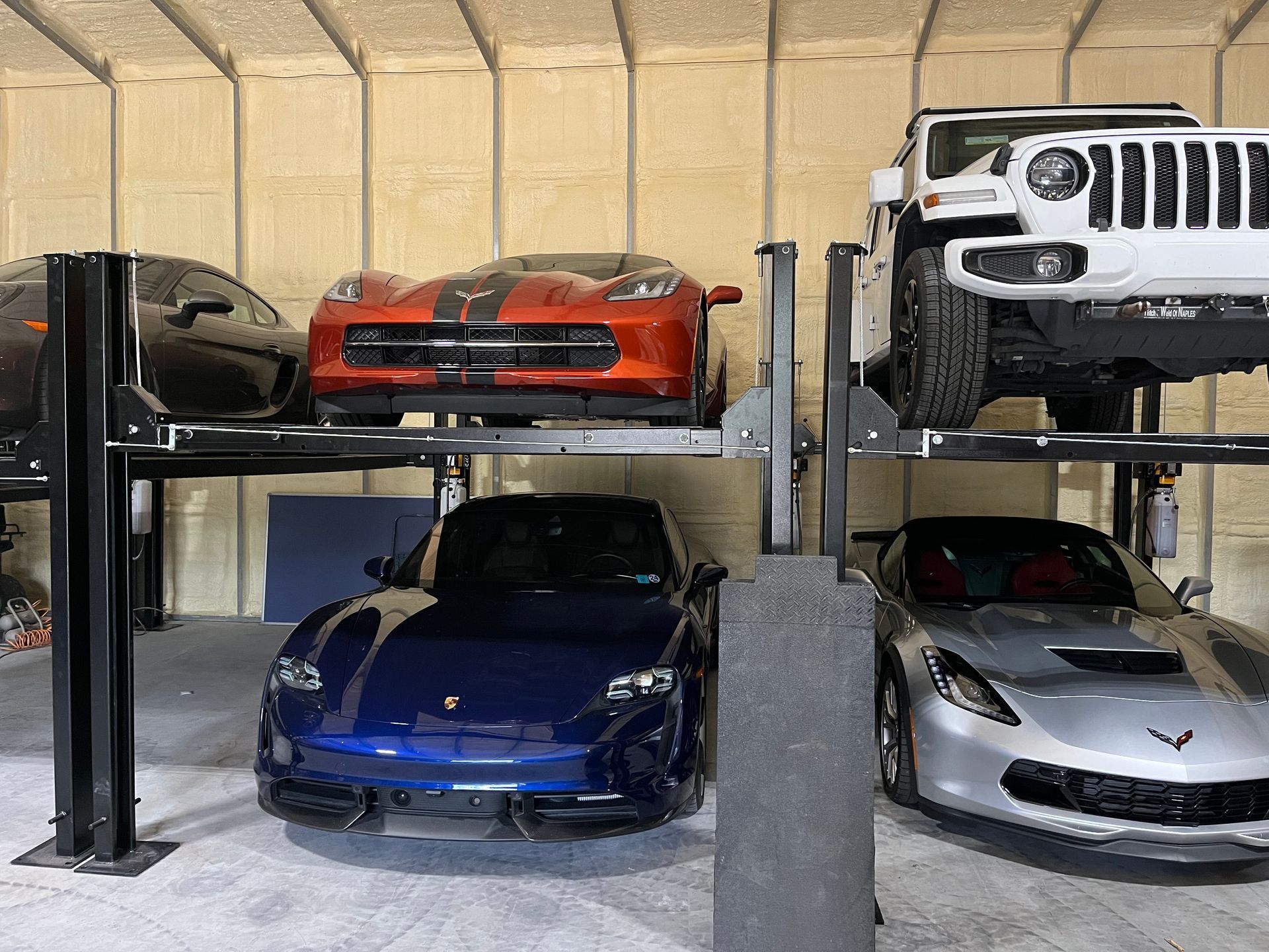 Four cars parked in a garage on two-level metal stacker lifts: an orange coupe, a white Jeep, a blue sports car, and a silver car.