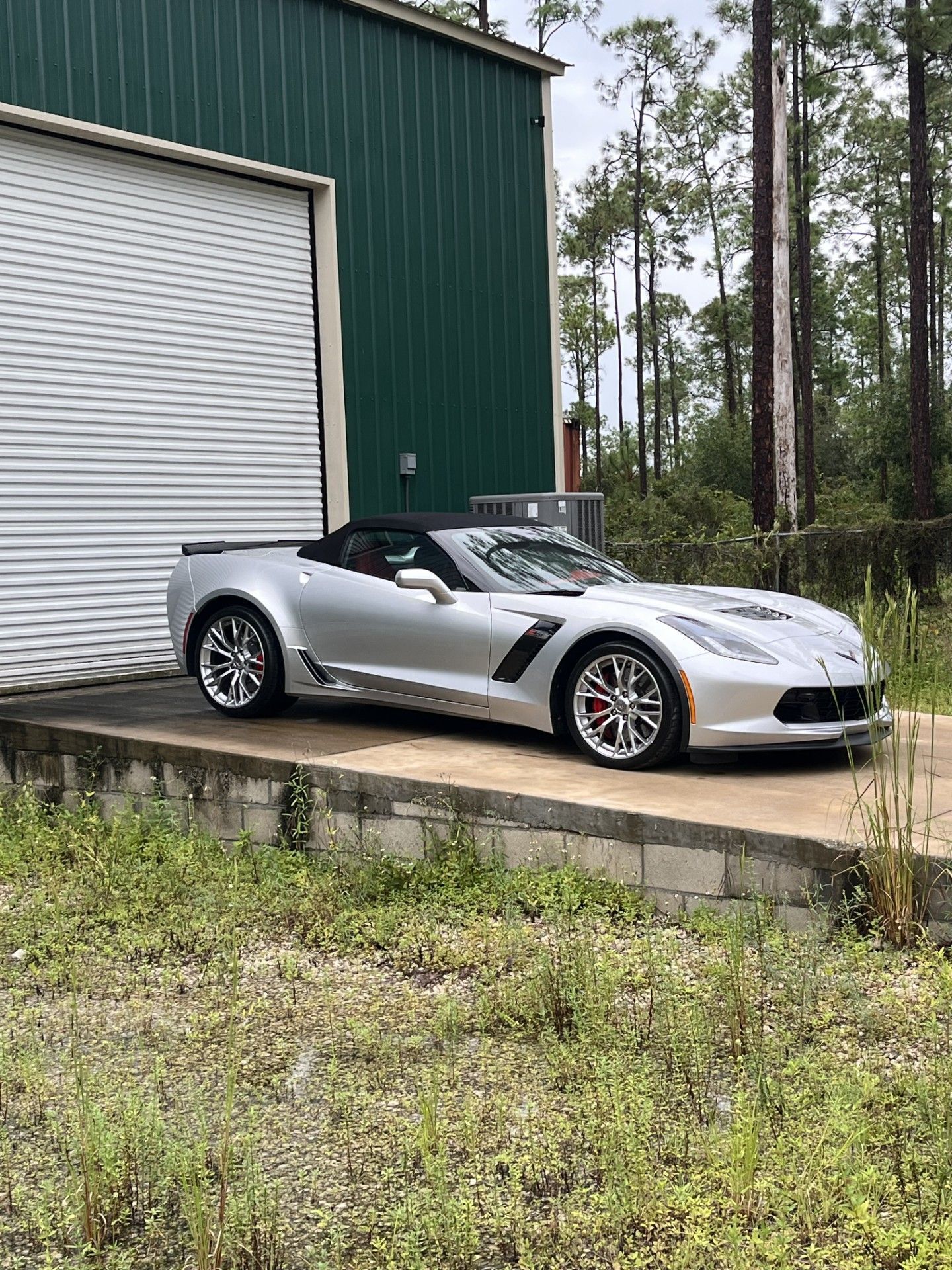 A silver Chevrolet Corvette convertible parked on a concrete ramp next to a green metal building.