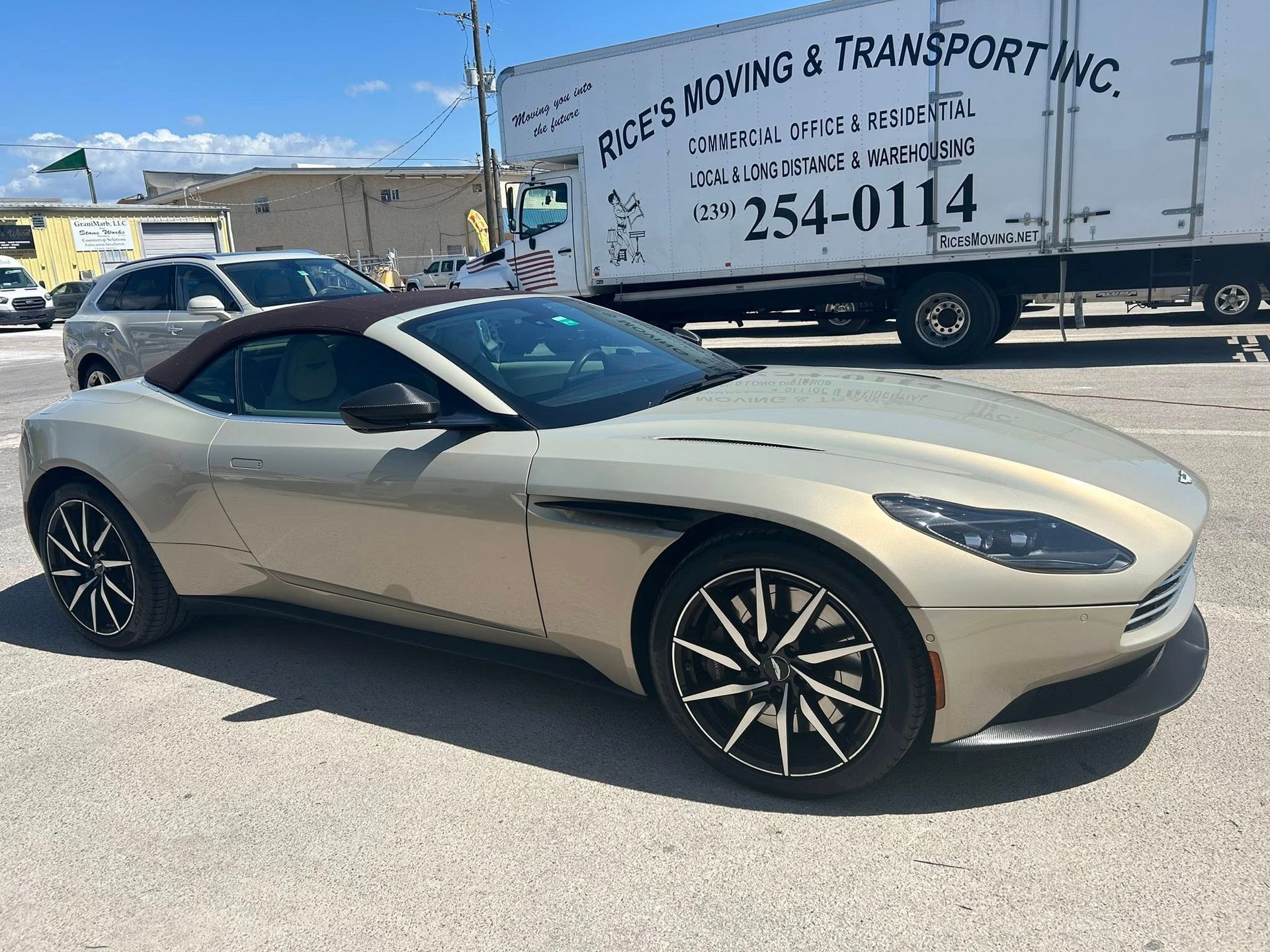A gold Aston Martin convertible parked in a lot next to a white moving truck.