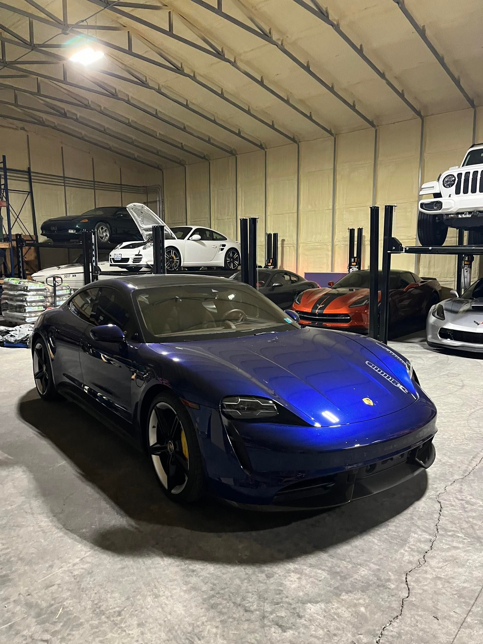 A shiny blue Porsche Taycan parked in a well-lit, spacious garage with several other luxury vehicles on car lifts.