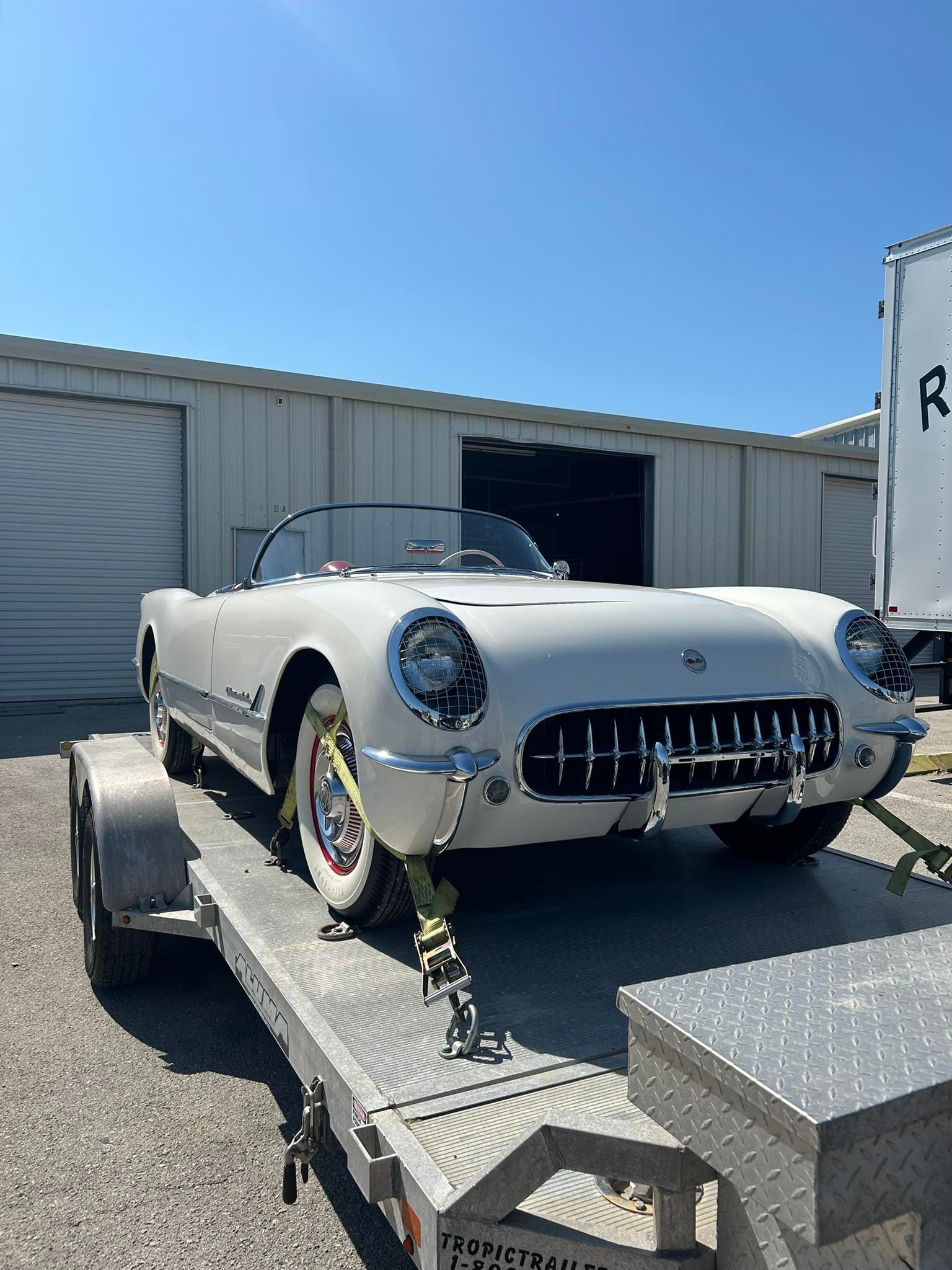 A vintage white Chevrolet Corvette convertible secured with yellow tie-down straps on a flatbed trailer.