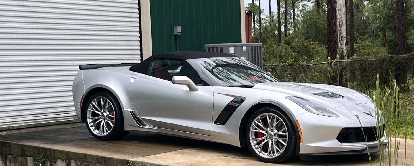 A silver Chevrolet Corvette convertible parked on a concrete driveway next to a building.