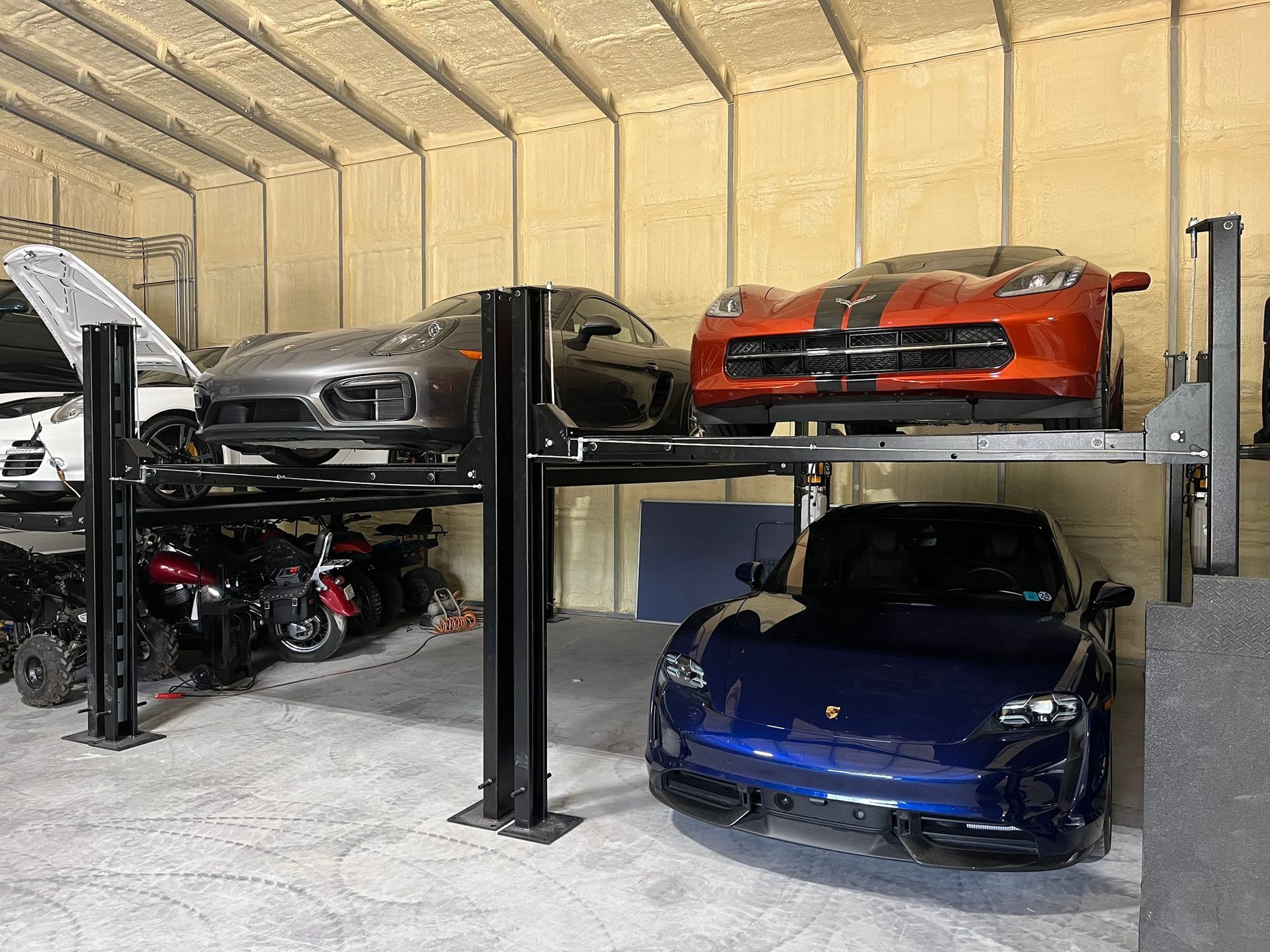 A garage with three sports cars parked, including a blue car below a two-tier lift holding gray and orange cars.