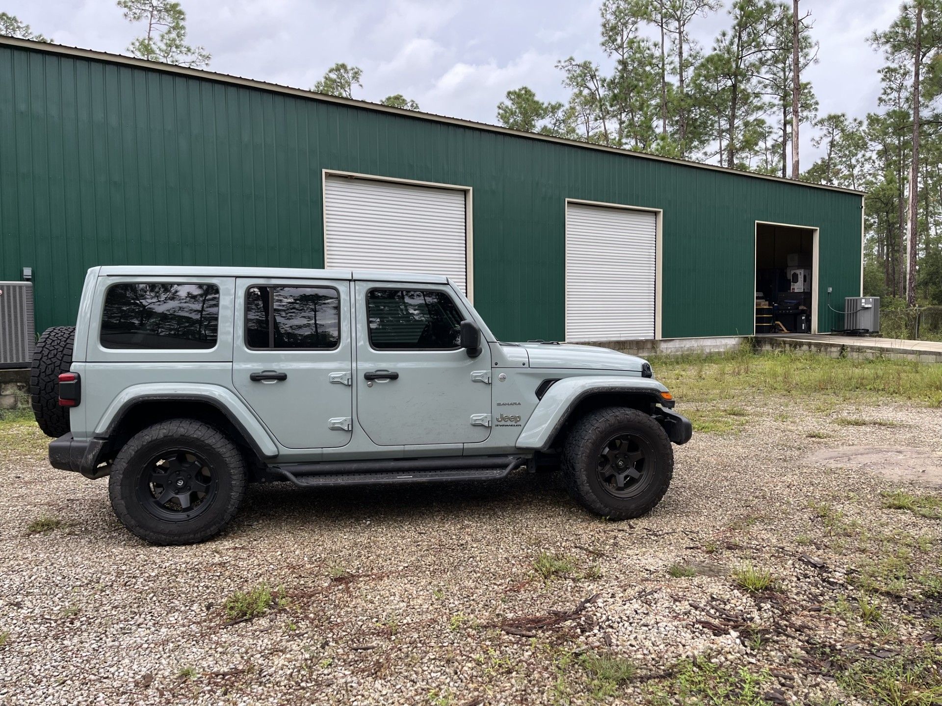 A light gray Jeep Wrangler parked on a gravel lot in front of a green metal industrial building.