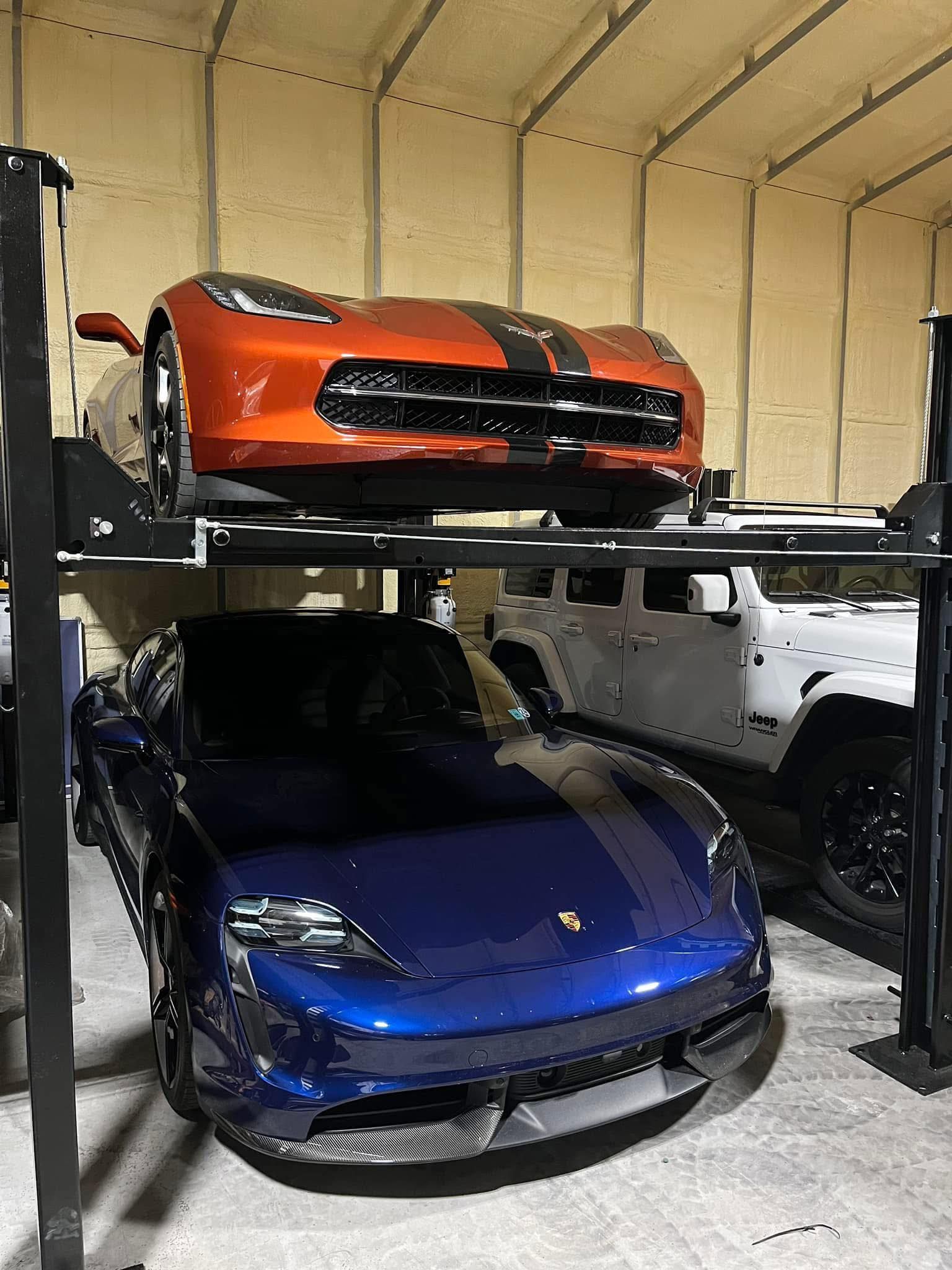 A blue Porsche Taycan parked in a garage below an orange Chevrolet Corvette on a vehicle lift, with a white Jeep nearby.