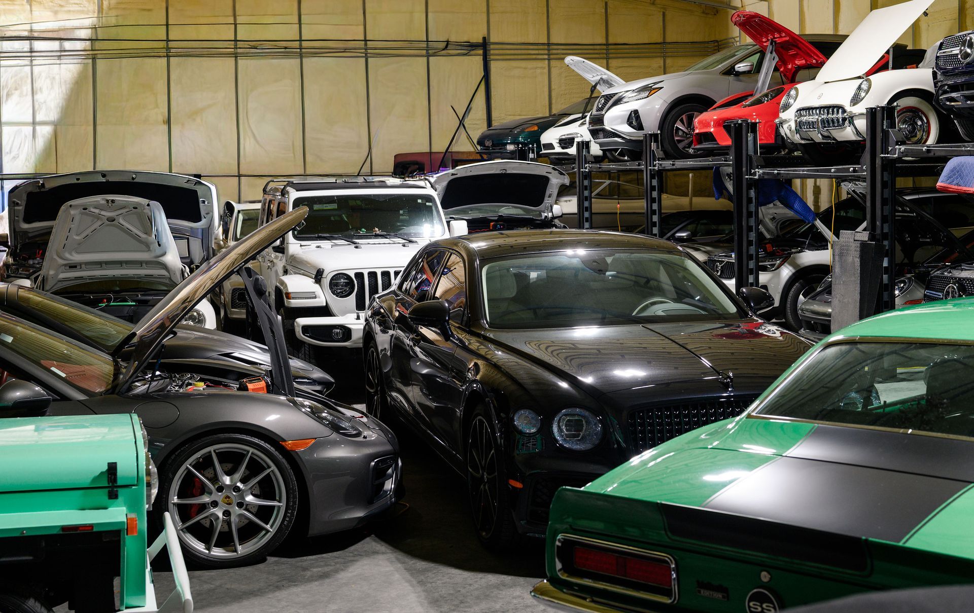 A crowded warehouse space filled with various vehicles, including a black Bentley, a white Jeep, and a green sports car.