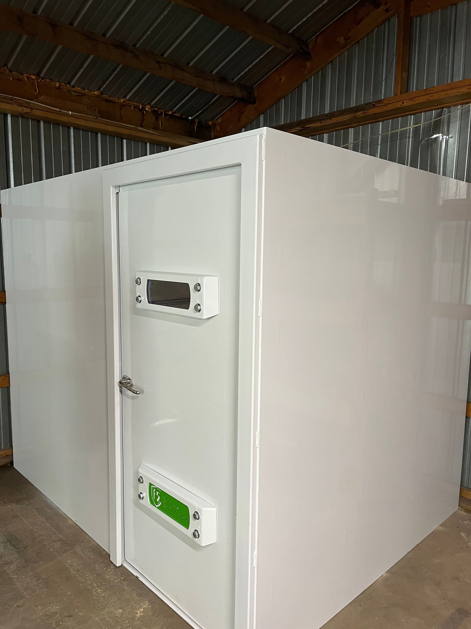 White walk-in cold room with a door and control panel inside a metal-roofed building