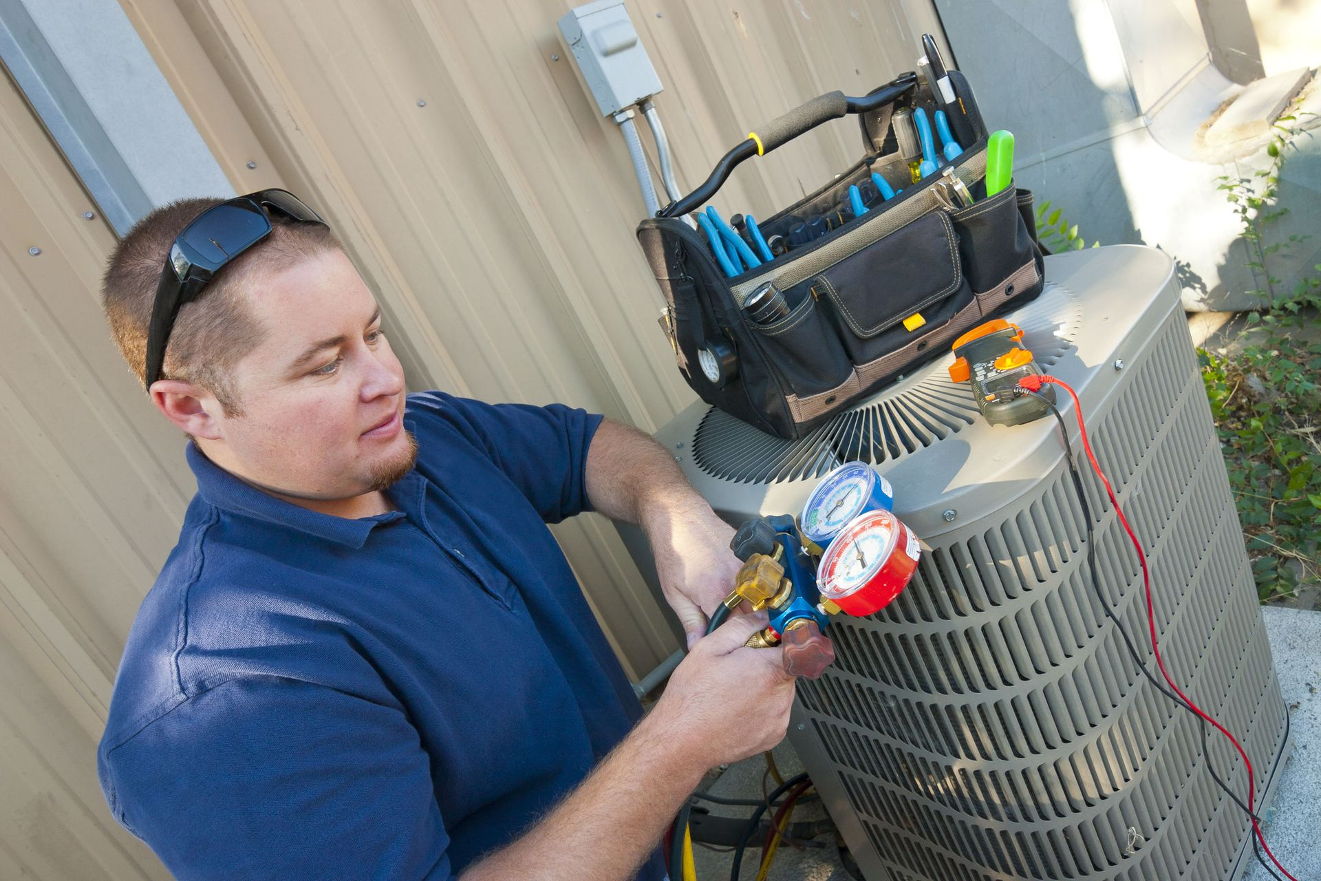 Technician checking an outdoor air conditioner unit with gauges and tools beside a beige wall