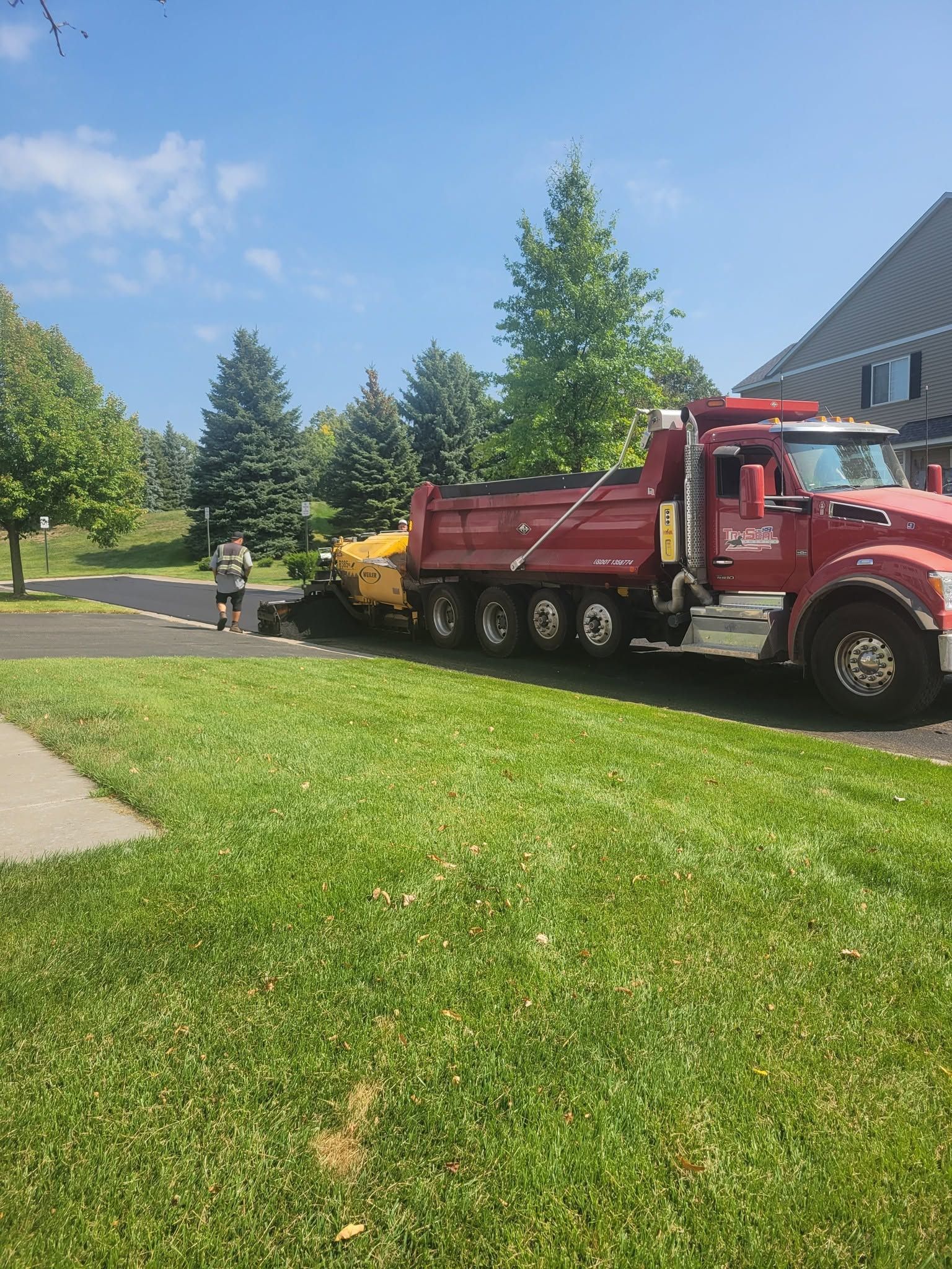 Red dump truck parked on a sunny lawn beside a sidewalk and trees.