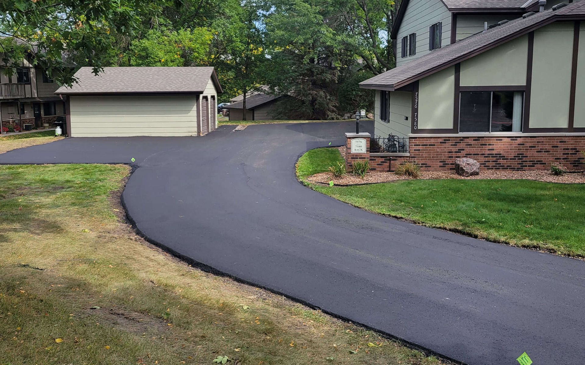 Freshly paved black driveway curving past a suburban house and garage, with green lawn and trees.