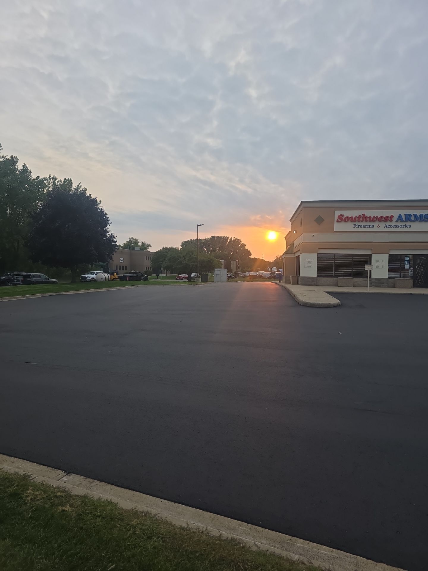 Sunset over an empty parking lot beside a storefront with a lit sign and trees on the left