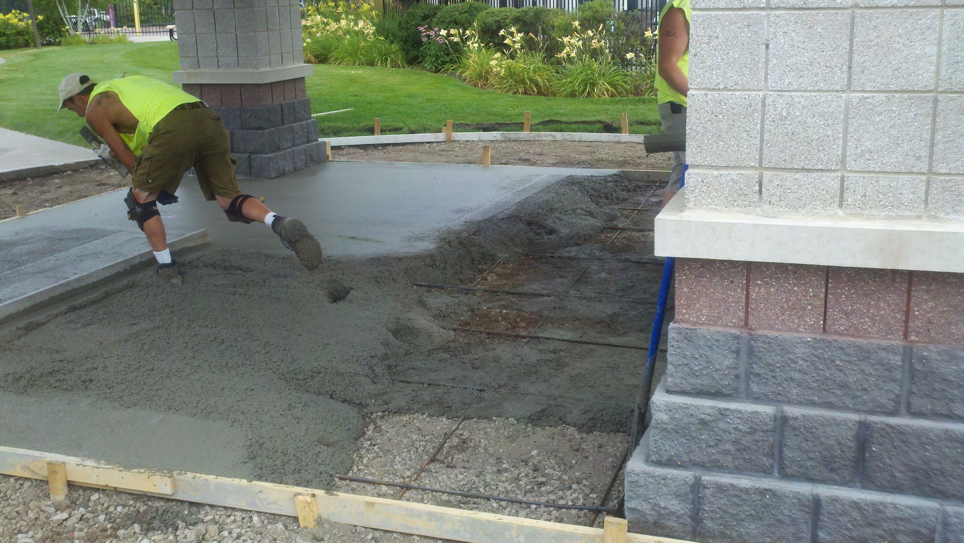 Man in a neon green shirt spreading fresh concrete beside a stone pavilion in a park