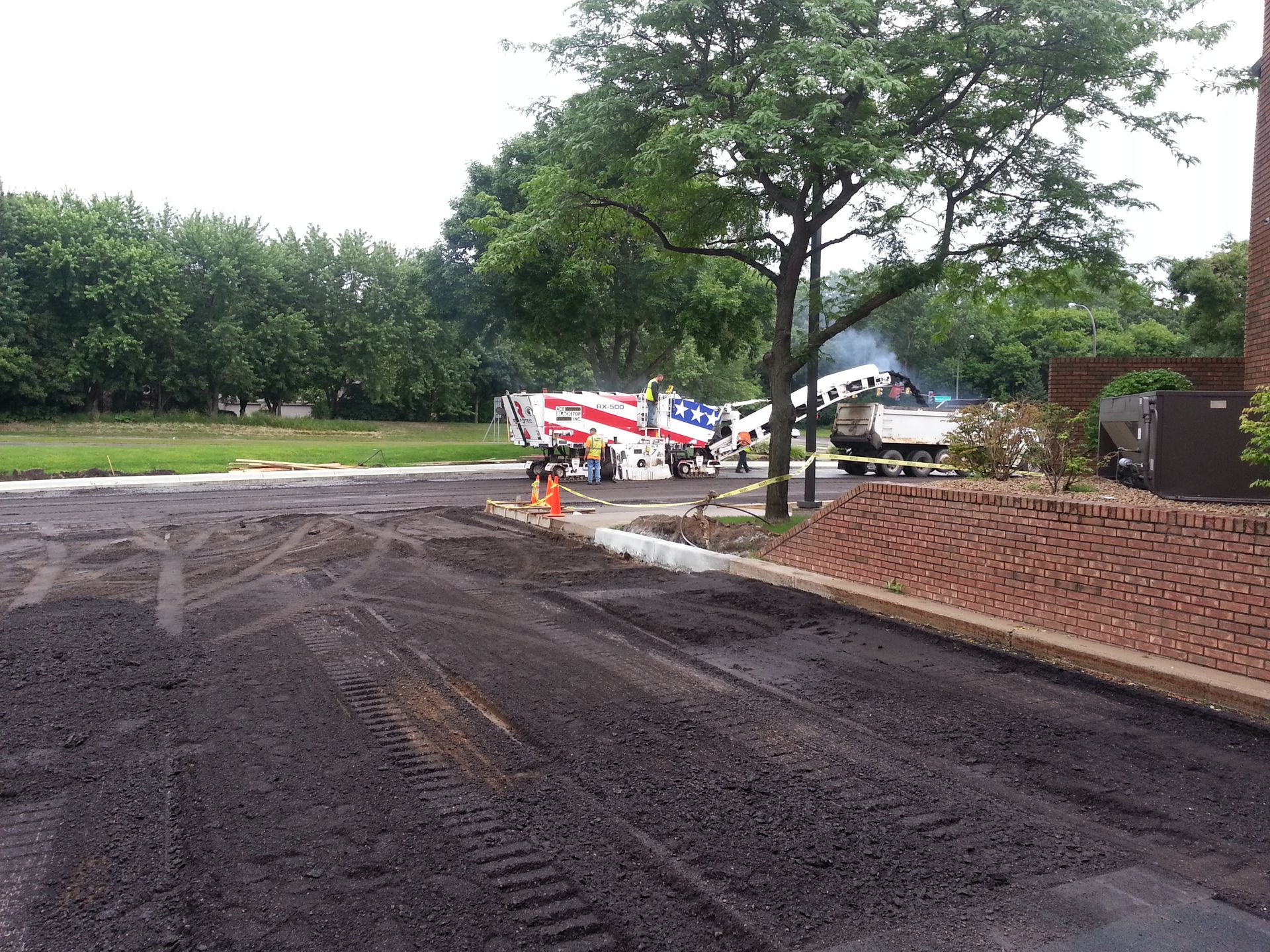 Roadwork scene with fresh asphalt, a tree-lined verge, and workers near equipment in the distance