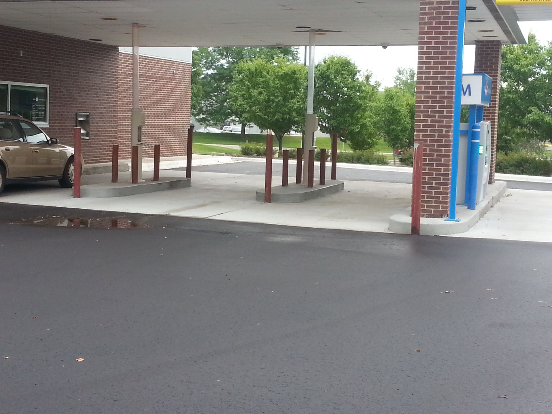 Drive-thru lane with concrete bollards, brick columns, parked car, and a blue parking meter kiosk under a canopy.