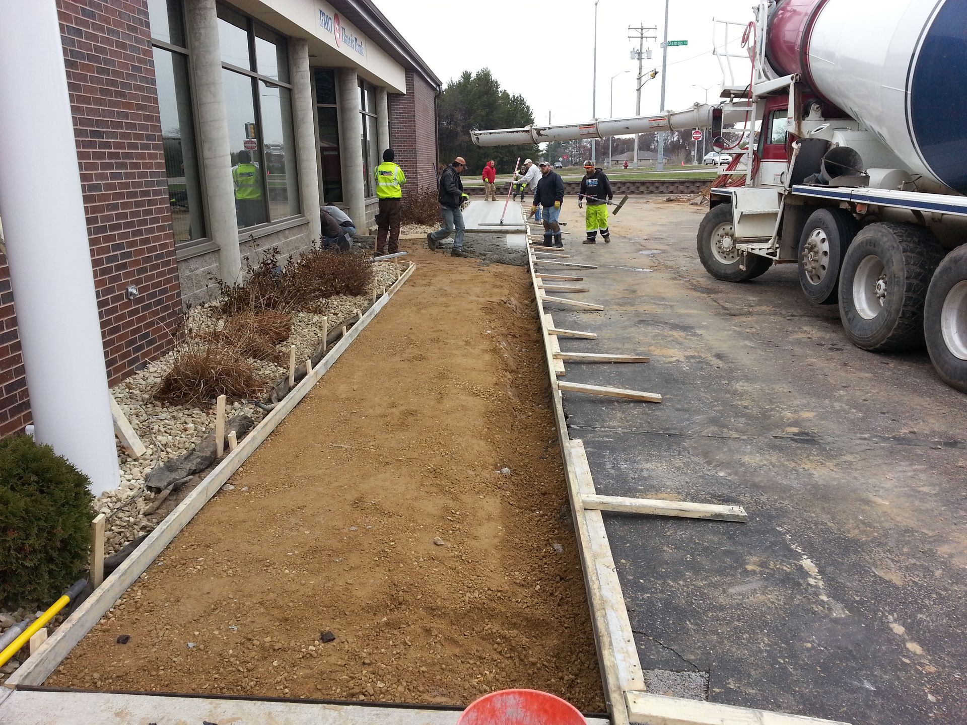 Workers beside a building pour and level dirt for a new landscaped strip next to a cement truck.