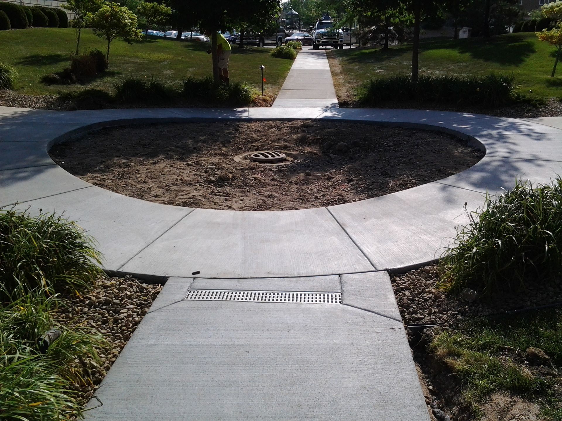 Circular concrete plaza with a central dirt planter and a walkway leading through a green park.