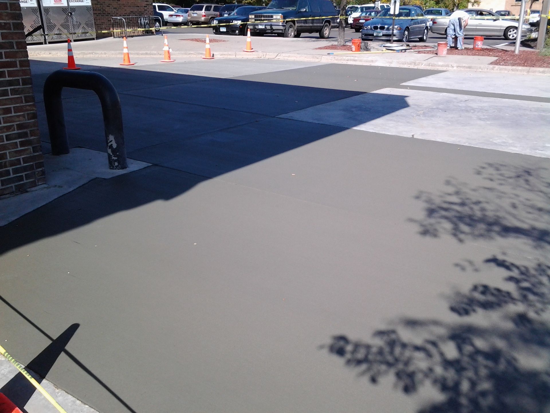 Concrete sidewalk beside a brick building, with parked cars and orange cones in a sunny lot.