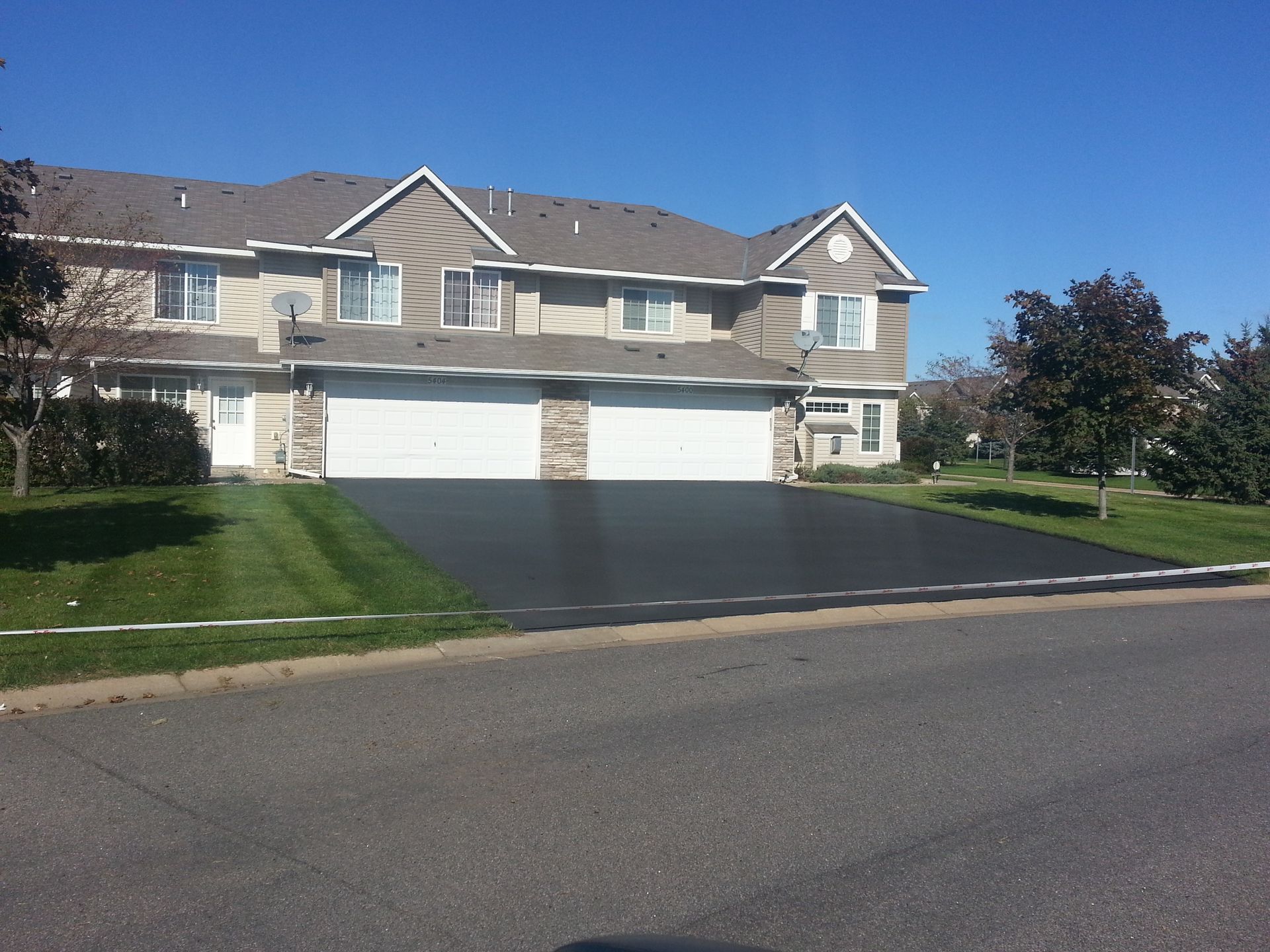 Suburban townhouse row with white garage doors, dark driveway, and green lawn under a clear blue sky