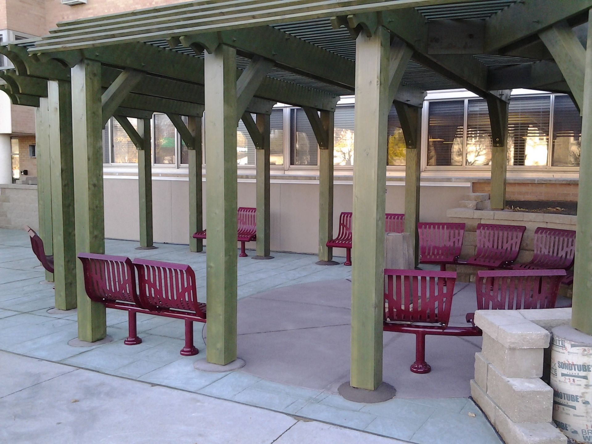 Covered outdoor seating area with red benches and green support posts on a paved plaza