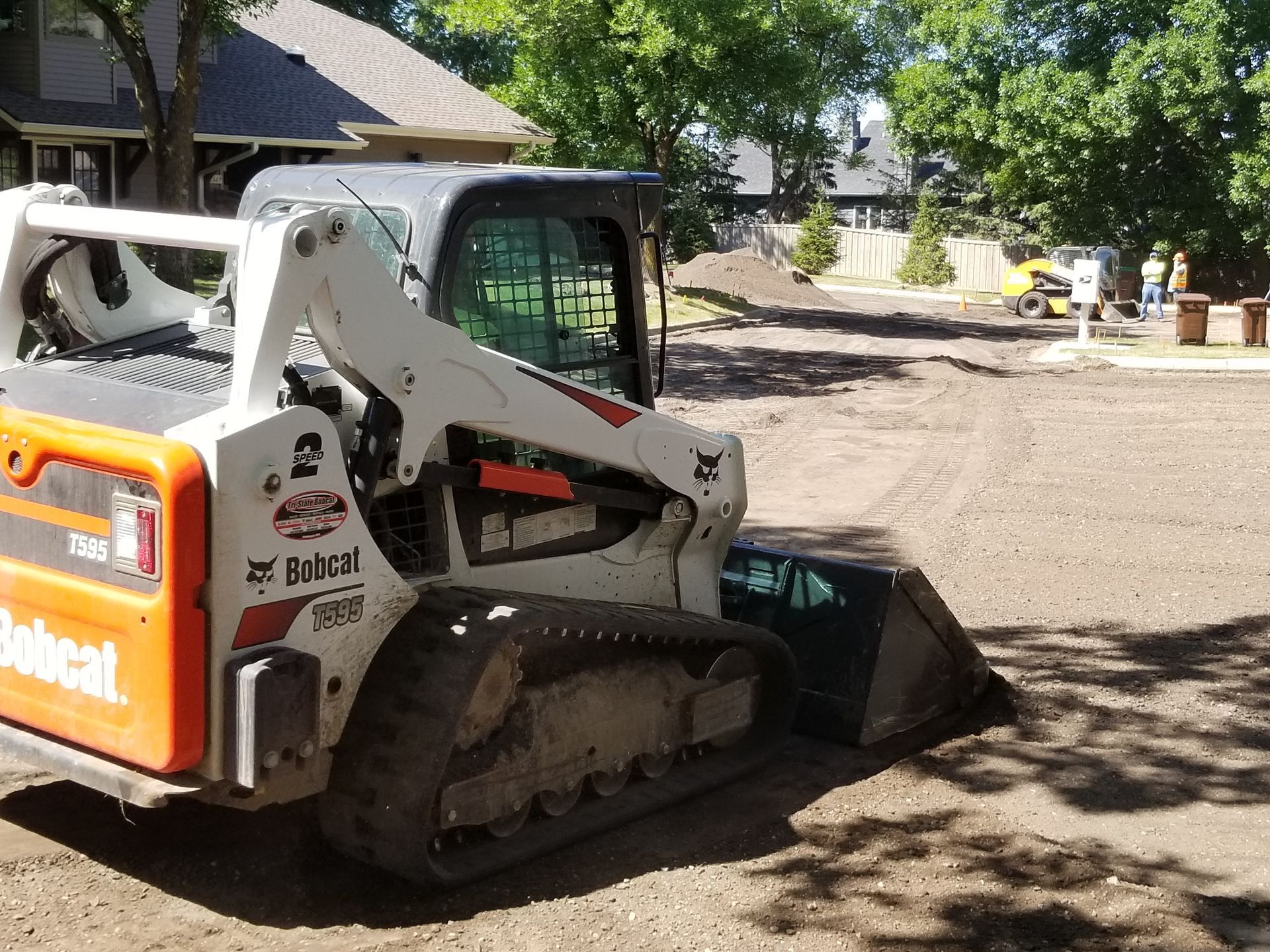 White Bobcat skid-steer loader on a gravel lot near trees and a house, bucket lowered in the foreground