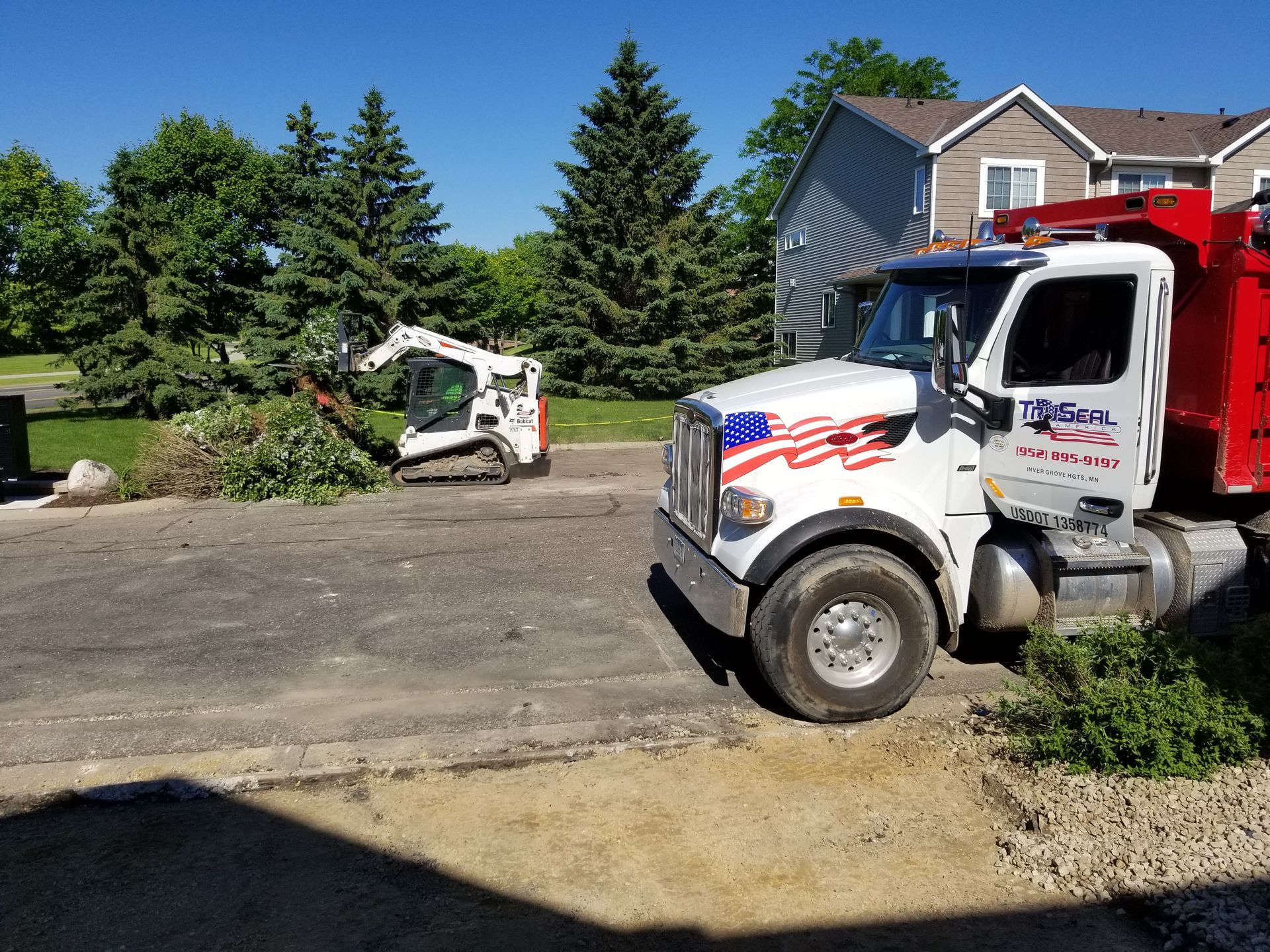 Two utility trucks and a skid steer clearing branches in a driveway beside a house.