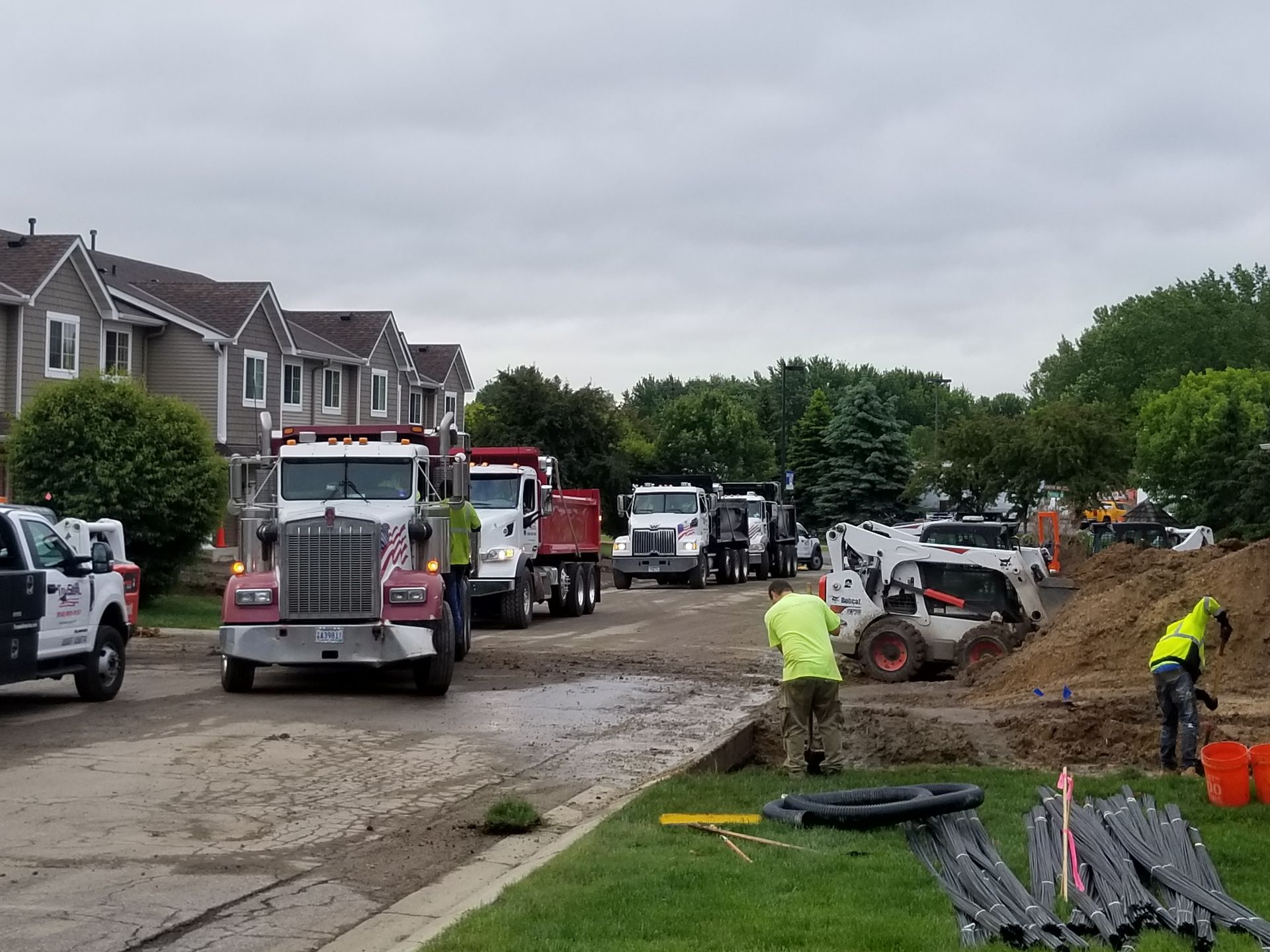 Utility trucks and workers at a muddy roadside construction site near houses under a cloudy sky