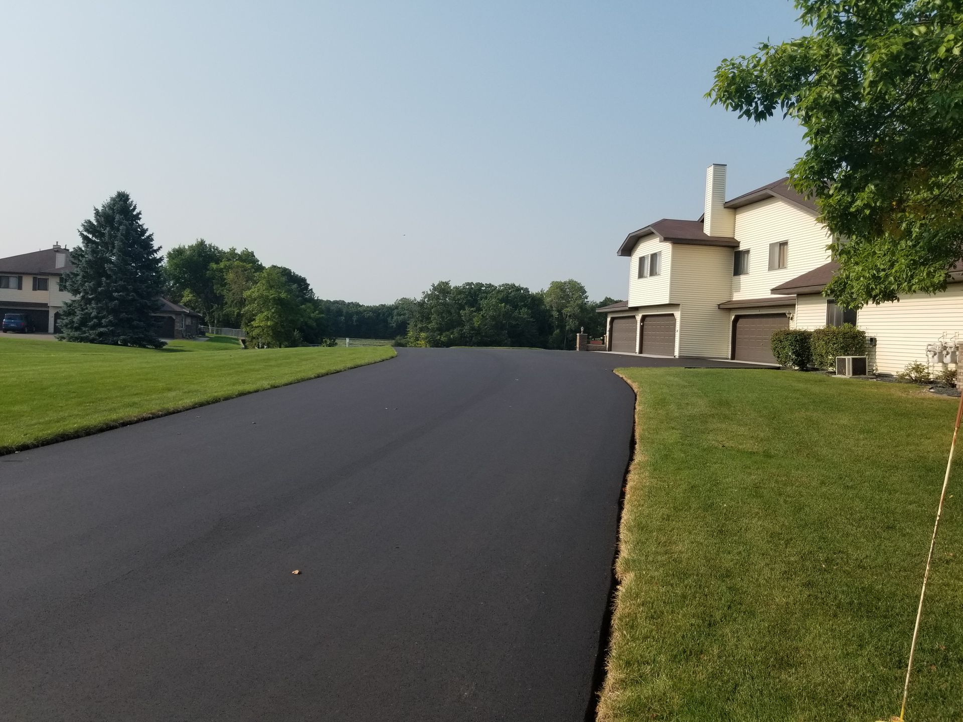 Quiet suburban street with fresh black asphalt, green lawns, and houses under a clear blue sky