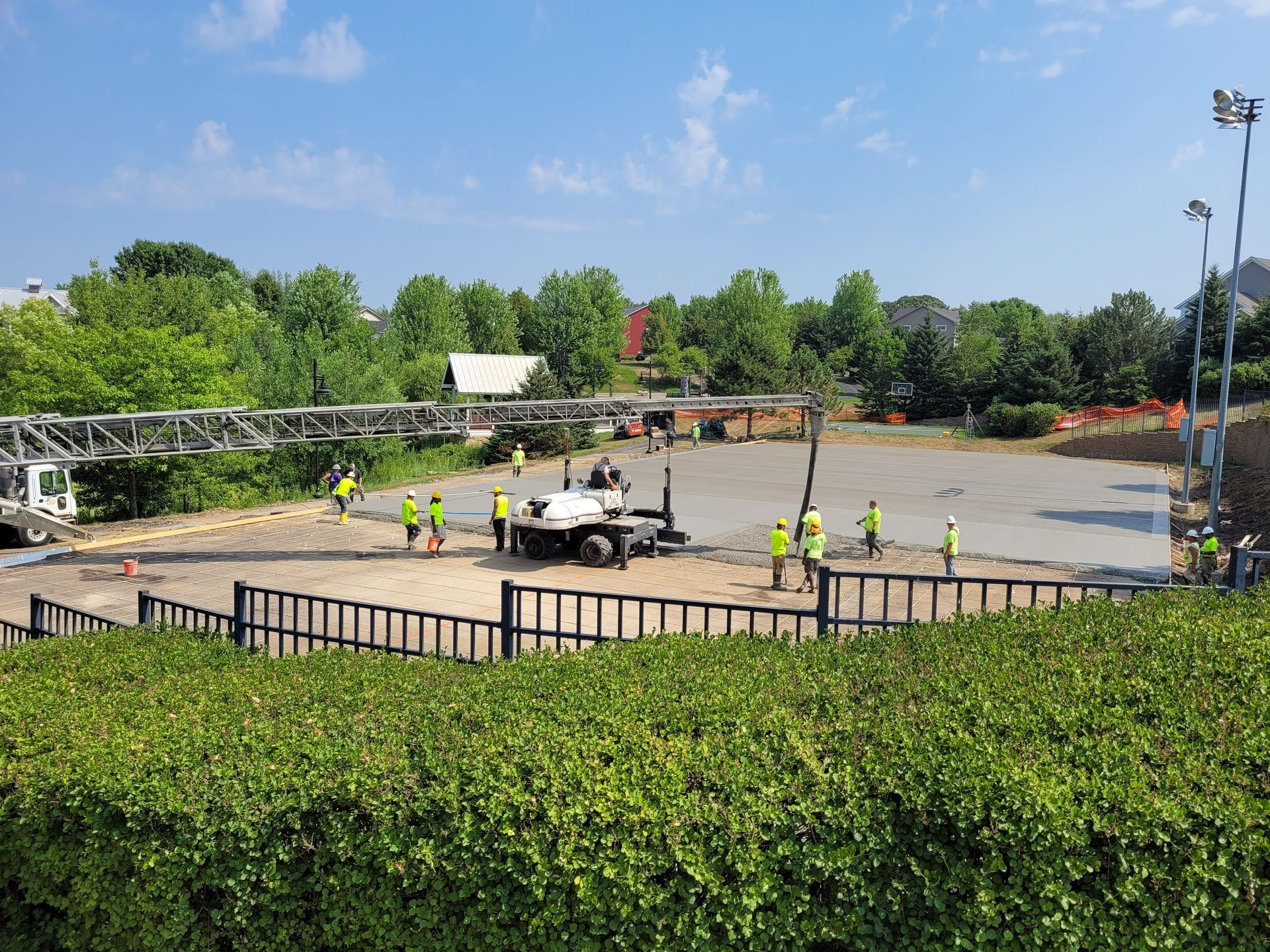 Workers repaving a parking lot beside a hedge, with a white truck and orange cones under a blue sky