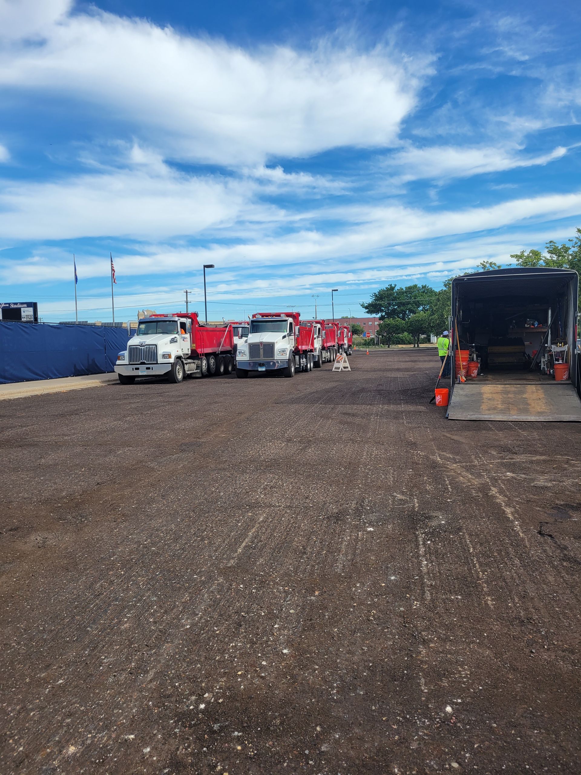 Gravel lot with parked work trucks and a trailer under a partly cloudy blue sky