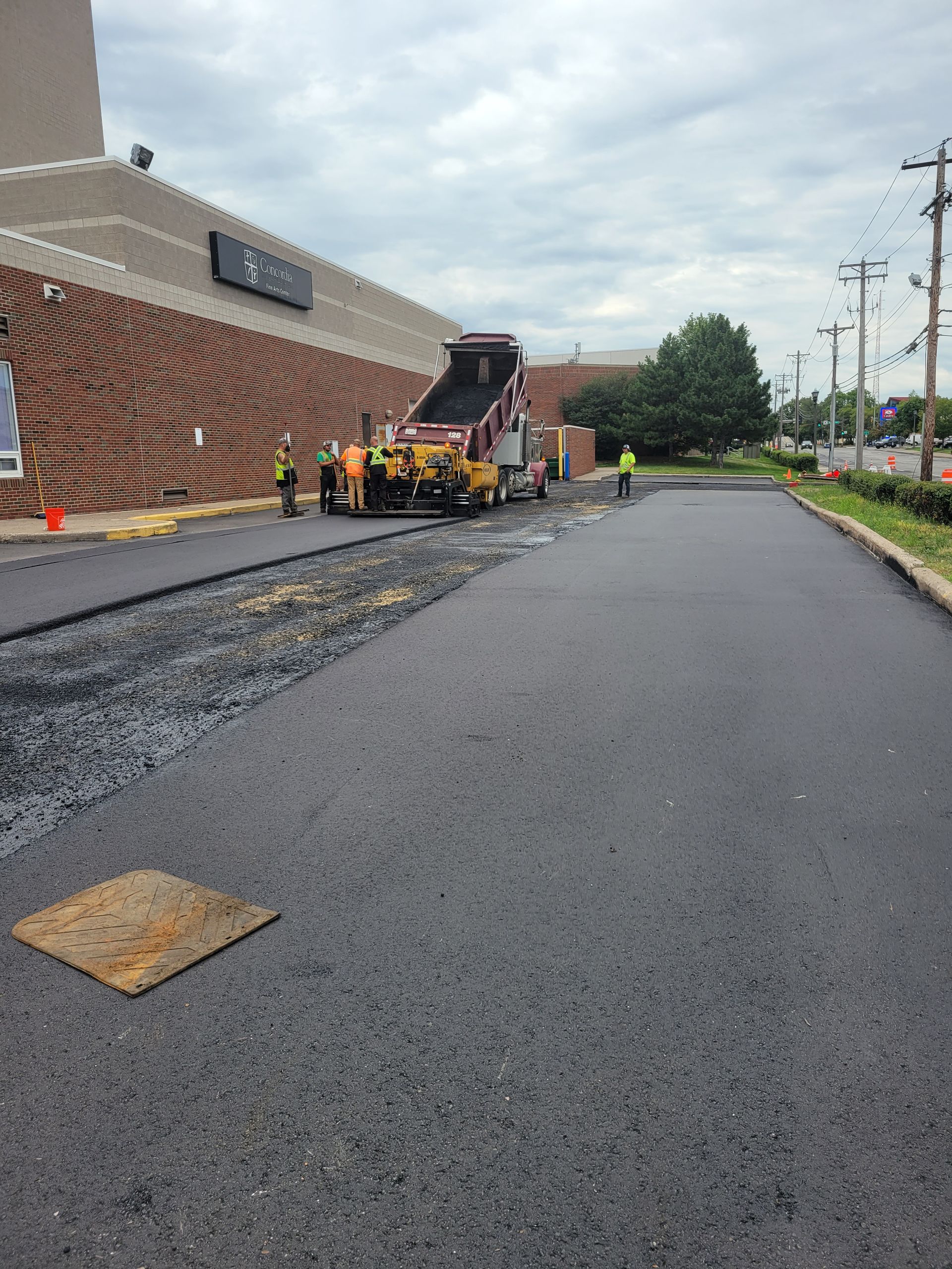 Roadwork crew paving a street beside a brick building, with asphalt machinery and cones under cloudy skies.