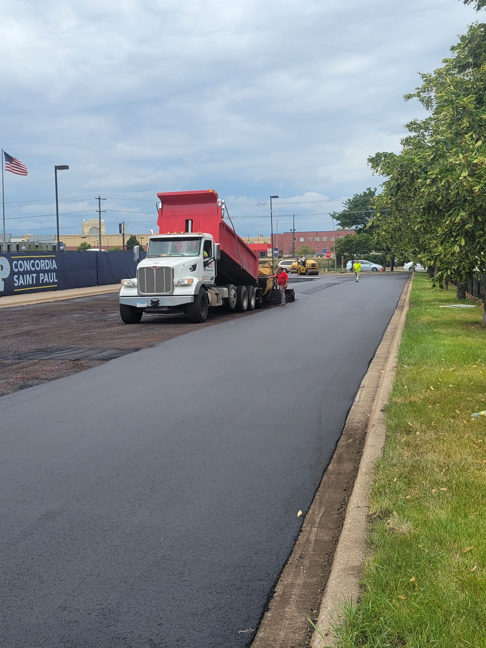 White truck hauling a red dump trailer on a freshly paved road beside a grassy verge under cloudy skies