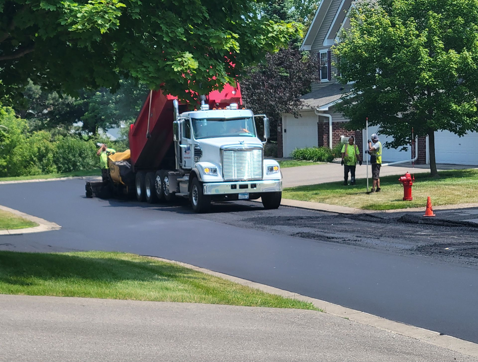 White truck hauling a red load on a residential street near workers and orange cones