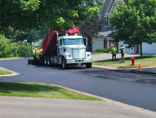 White dump truck unloading a red-covered load on a suburban street near a house and orange cones.