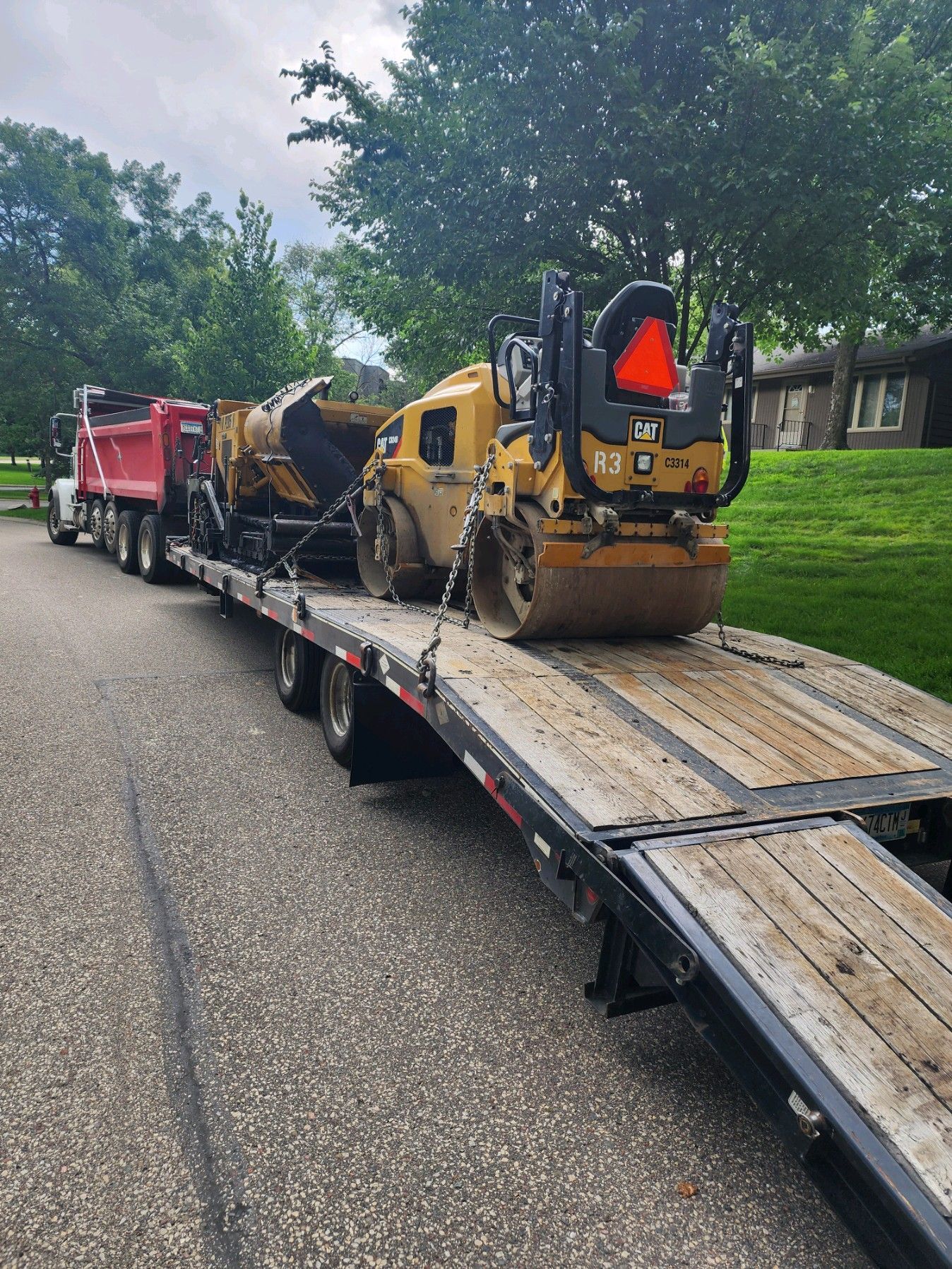 Yellow construction roller on a flatbed trailer with an orange warning sign, parked on a tree-lined road.
