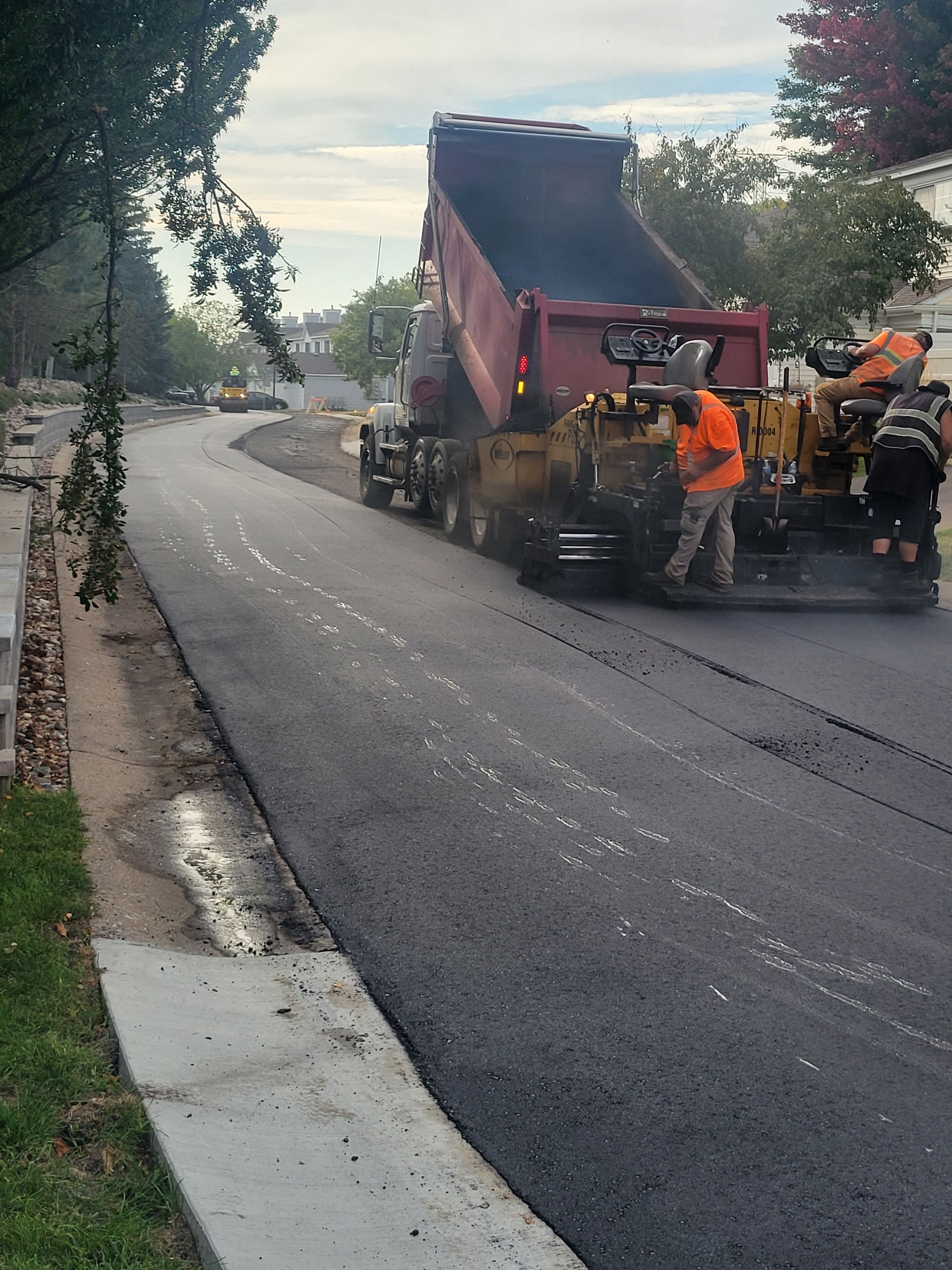 Road paving crew spreads fresh asphalt on a curved street beside a sidewalk and trees.