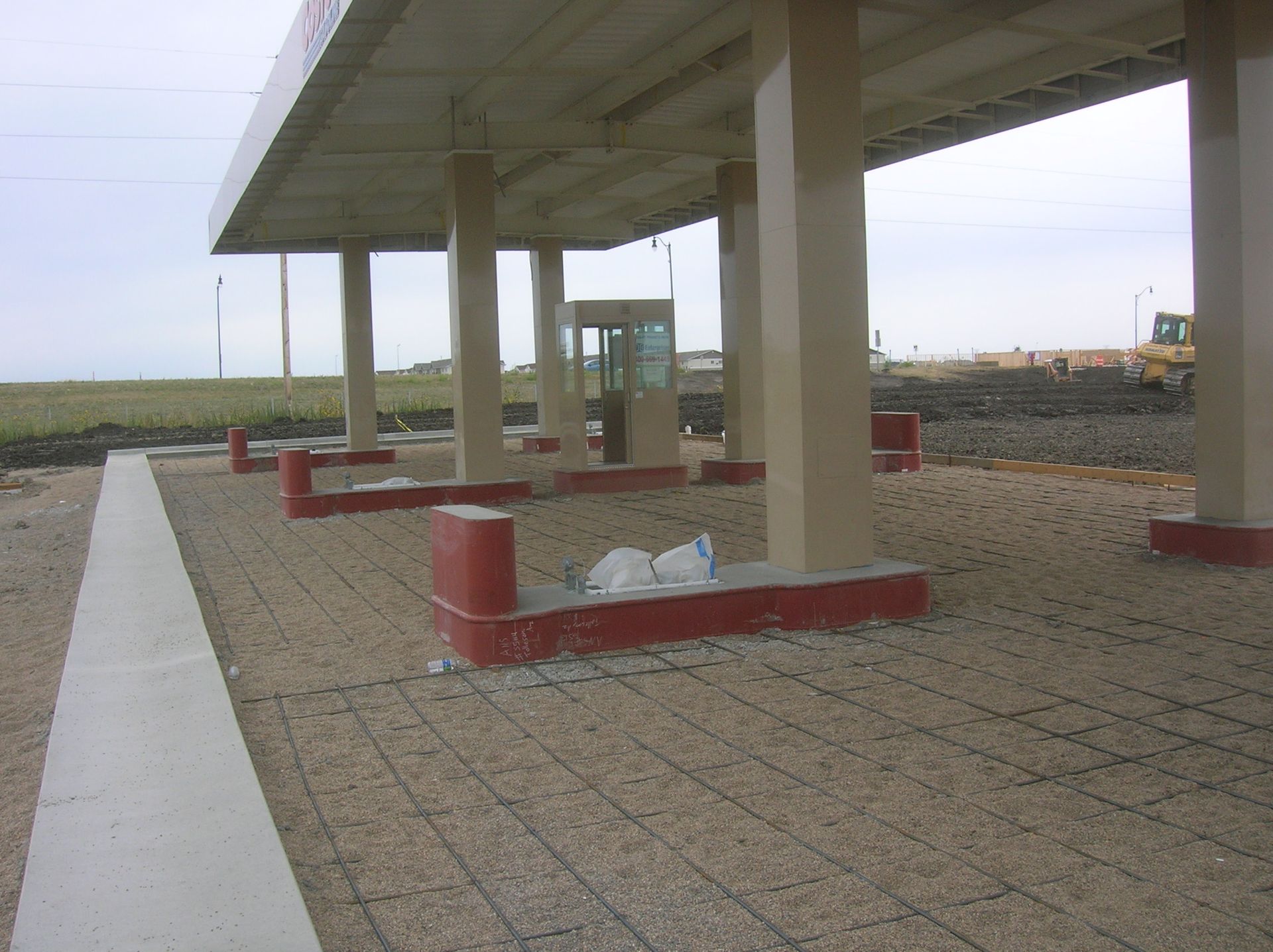 Concrete underpass with red barriers and support columns beside a tiled walkway in an open, sandy area