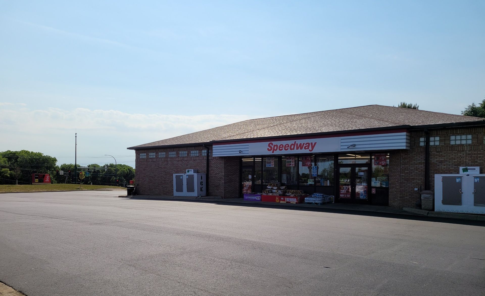 Small brick convenience store with a white sign and large parking lot under a blue sky
