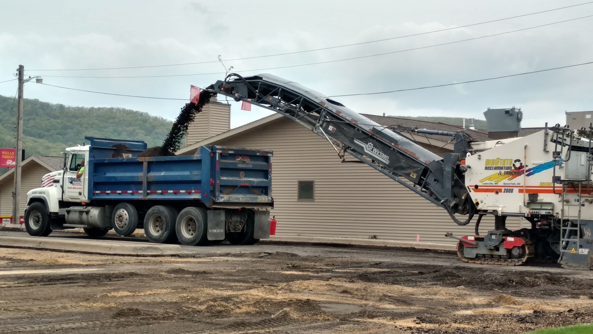 Road milling machine unloading asphalt into a blue dump truck on a roadside construction site