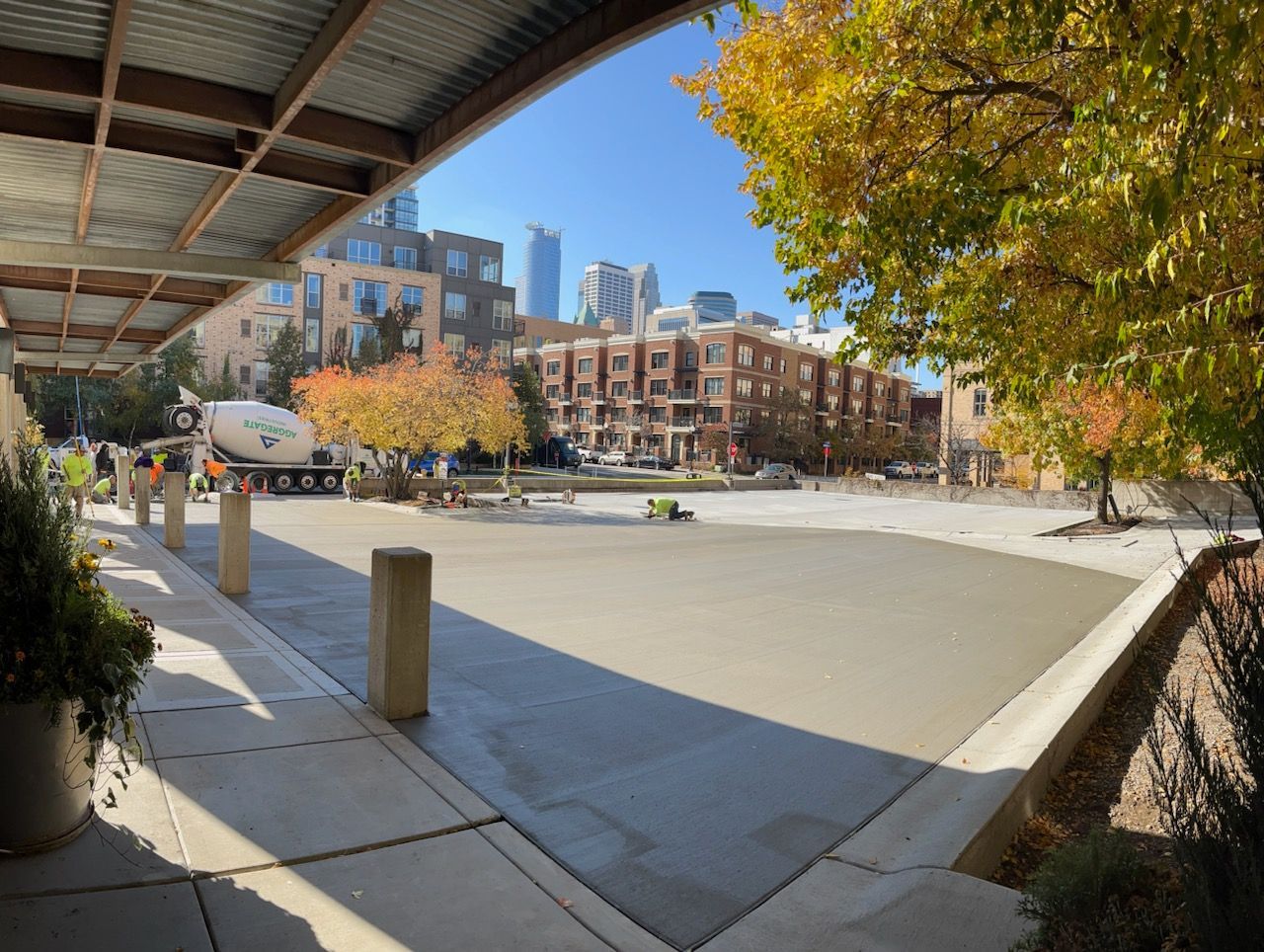 Sunny urban plaza with trees, a covered walkway, and brick buildings under a blue sky