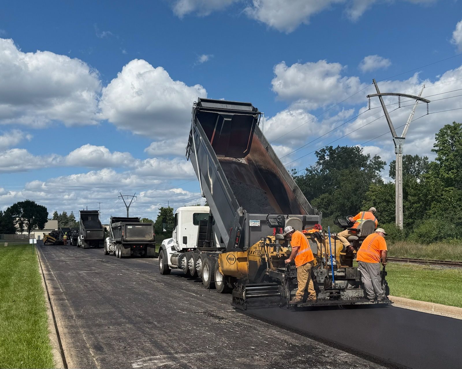 Road crew paving a highway, with dump trucks, an asphalt paver, and workers in orange safety gear