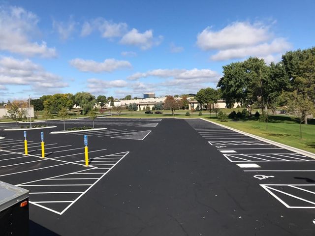 Empty parking lot with fresh black asphalt, white lines, yellow bollards, and trees under a blue sky.