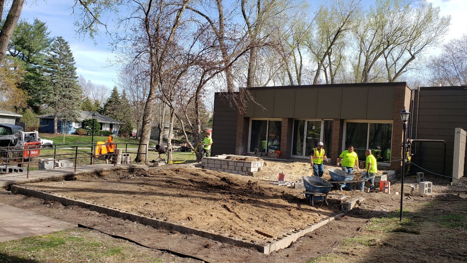 Workers in neon vests build a foundation beside a small brown building in a wooded park.