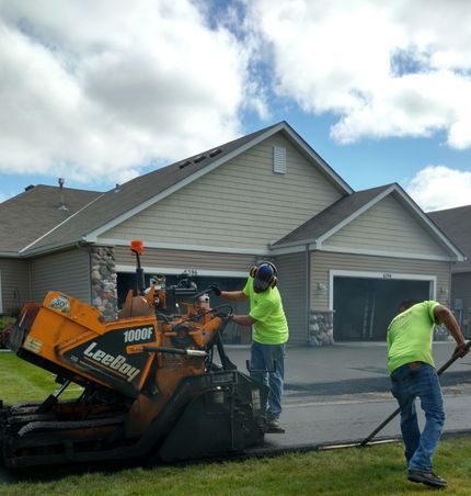 Workers operating an orange asphalt machine on a residential driveway beside a suburban house.