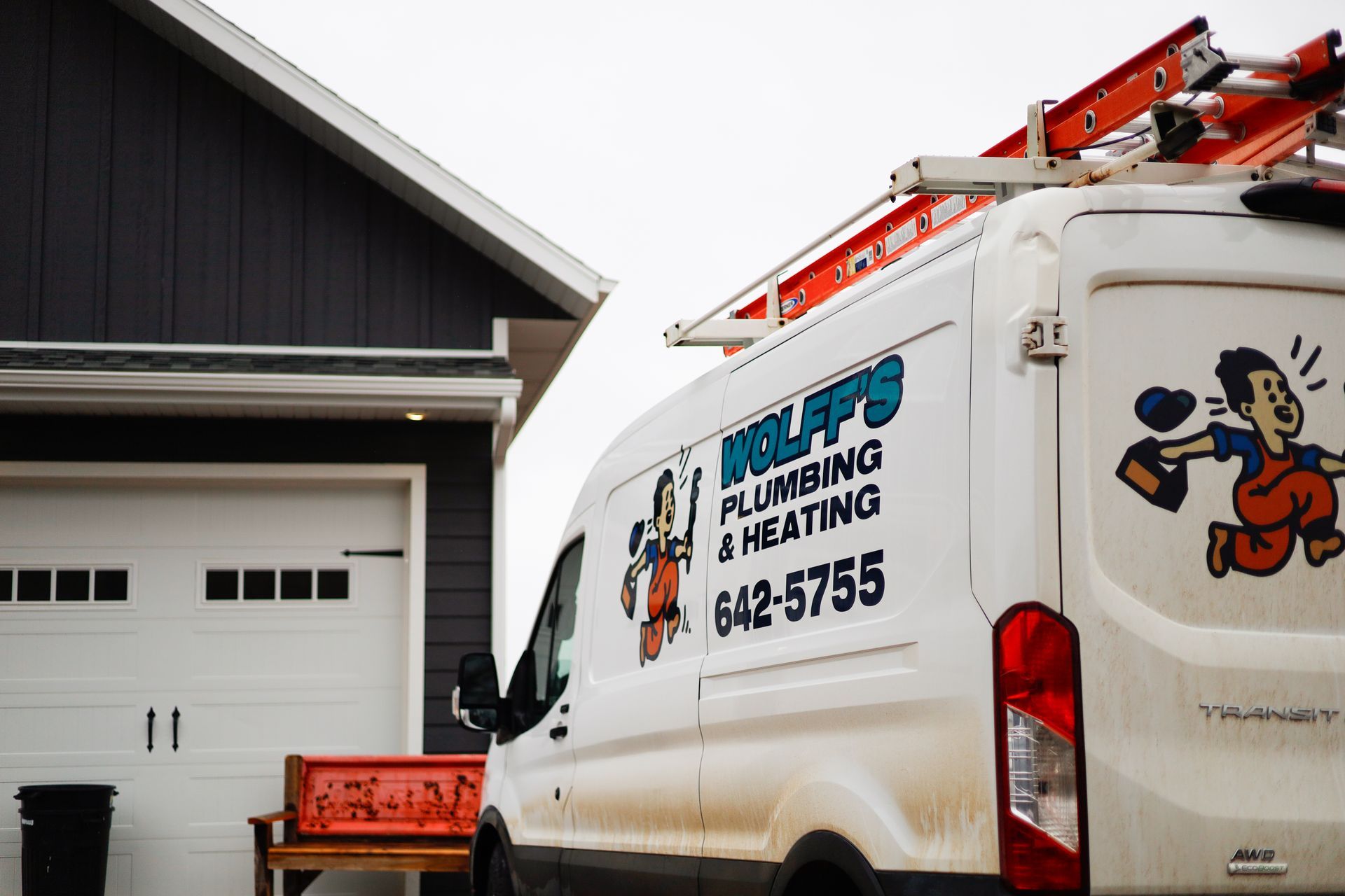White plumbing van parked near a house, ladder on top. Van has business name and cartoon plumber.