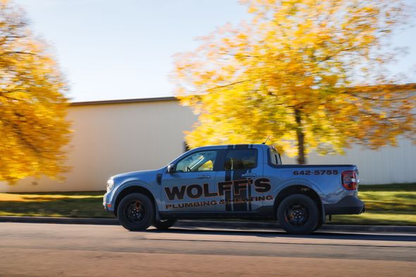 Blue Wolfe's Plumbing truck drives down a street in front of a building and trees with yellow leaves.