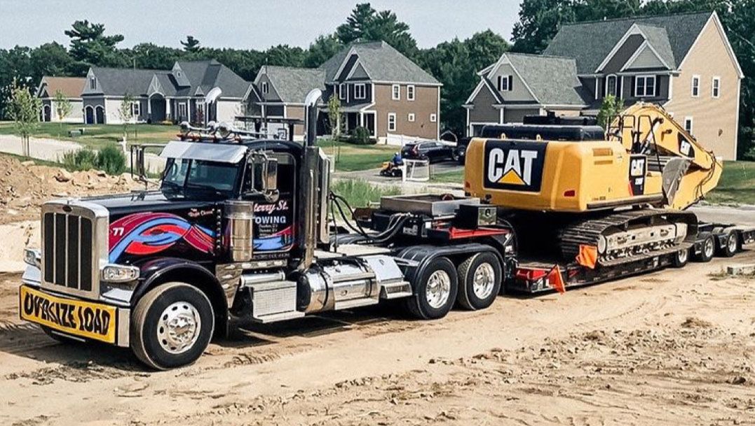 Blue semi truck hauling a yellow CAT excavator on a flatbed trailer at a construction site.