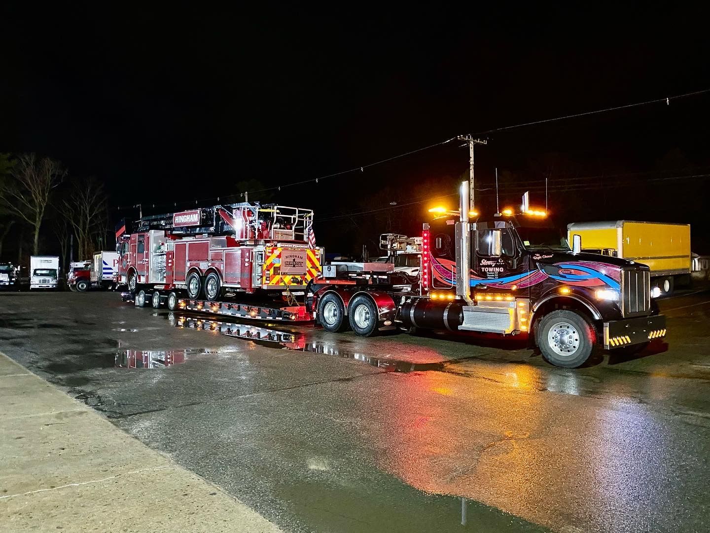 Fire trucks and a tow truck lined up on a wet road at night, lights reflecting on the pavement.