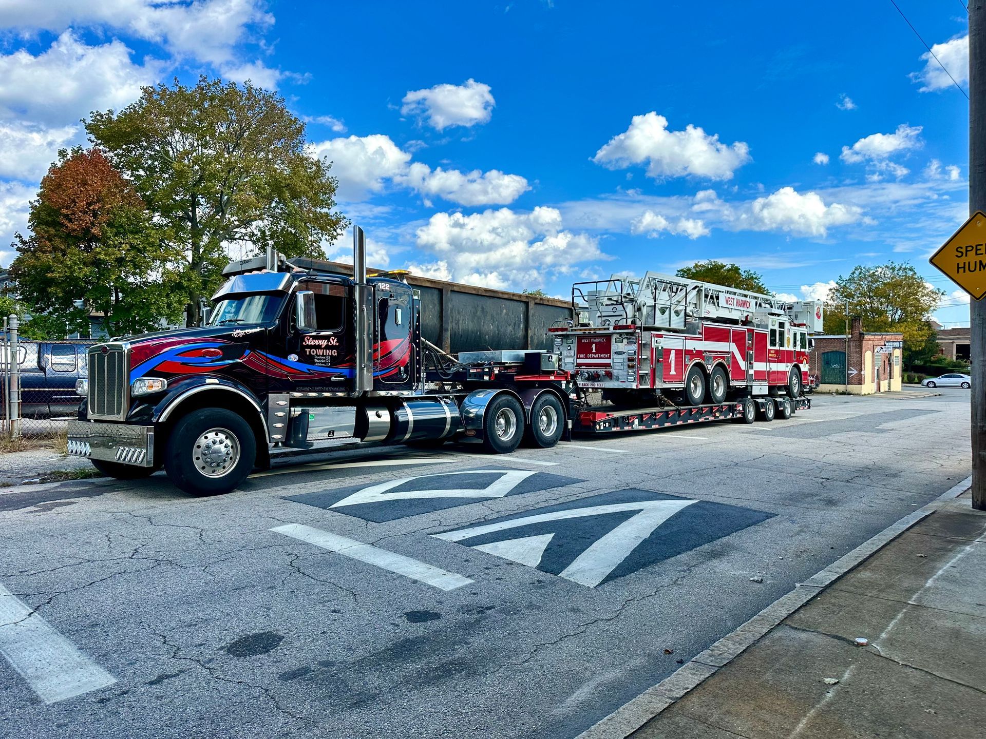 Large black and red semi-truck hauling a red fire engine on a road under a bright blue sky.