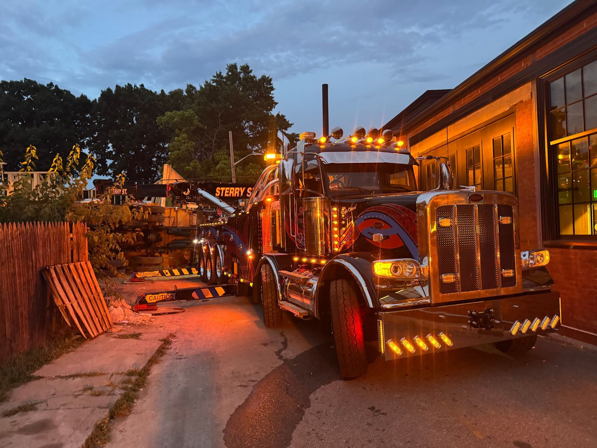 Tow truck parked at dusk beside a building, with orange lights glowing and a wooded background.